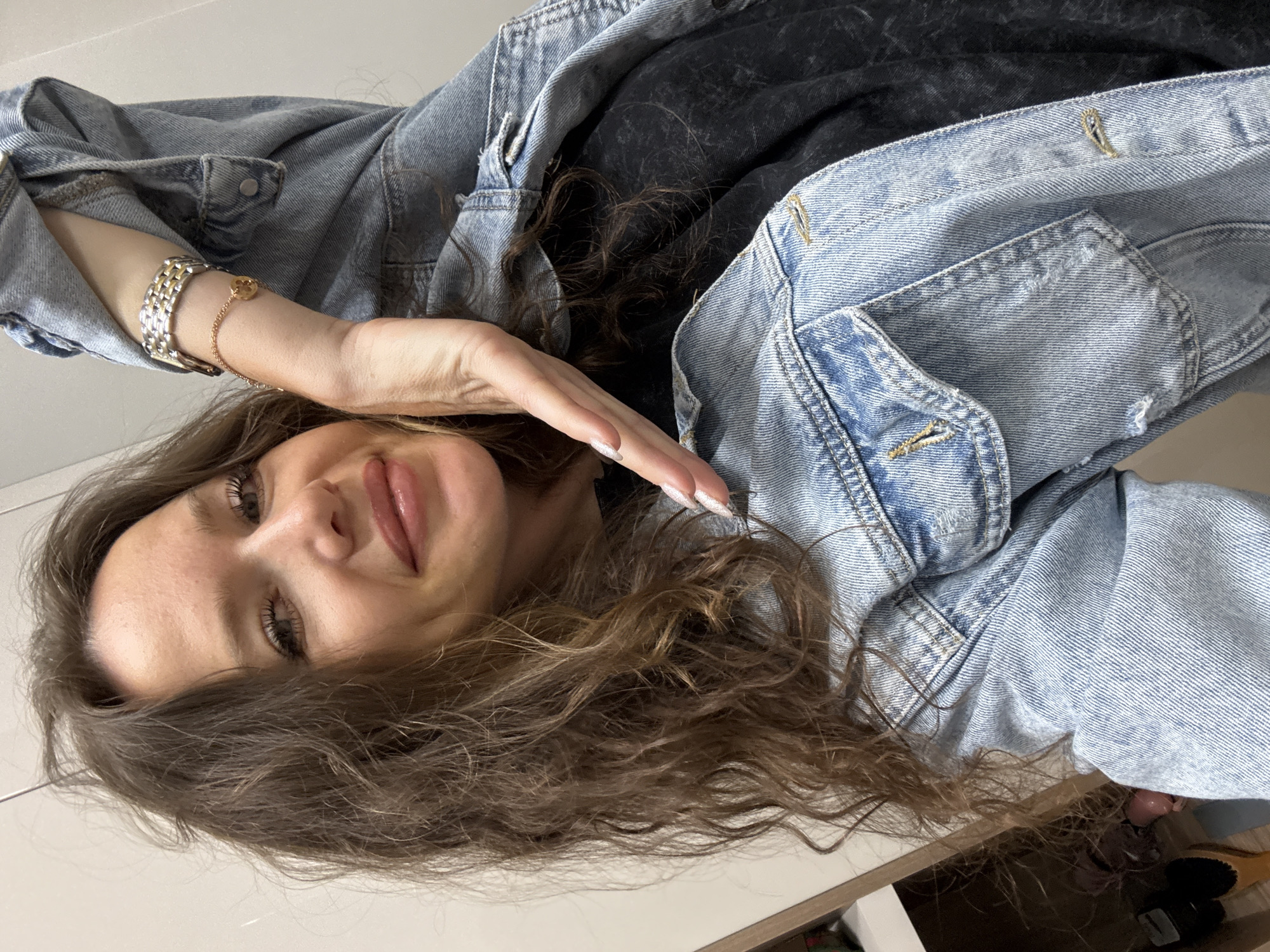 A young woman with long, wavy brown hair is posing for a selfie in what appears to be an indoor setting, possibly a closet or dressing area in Dolgoprudny, Russia. She is wearing a light-wash denim jacket over a dark t-shirt and has her left hand resting on her cheek, with her fingers extended upwards towards her temple. She is also wearing a silver and gold link bracelet on her left wrist and a delicate rose gold chain bracelet with a small charm. The background shows a pale wall and the edge of a shelf unit with some items visible on it, including what looks like a hairbrush and footwear. The lighting suggests an artificial indoor environment.