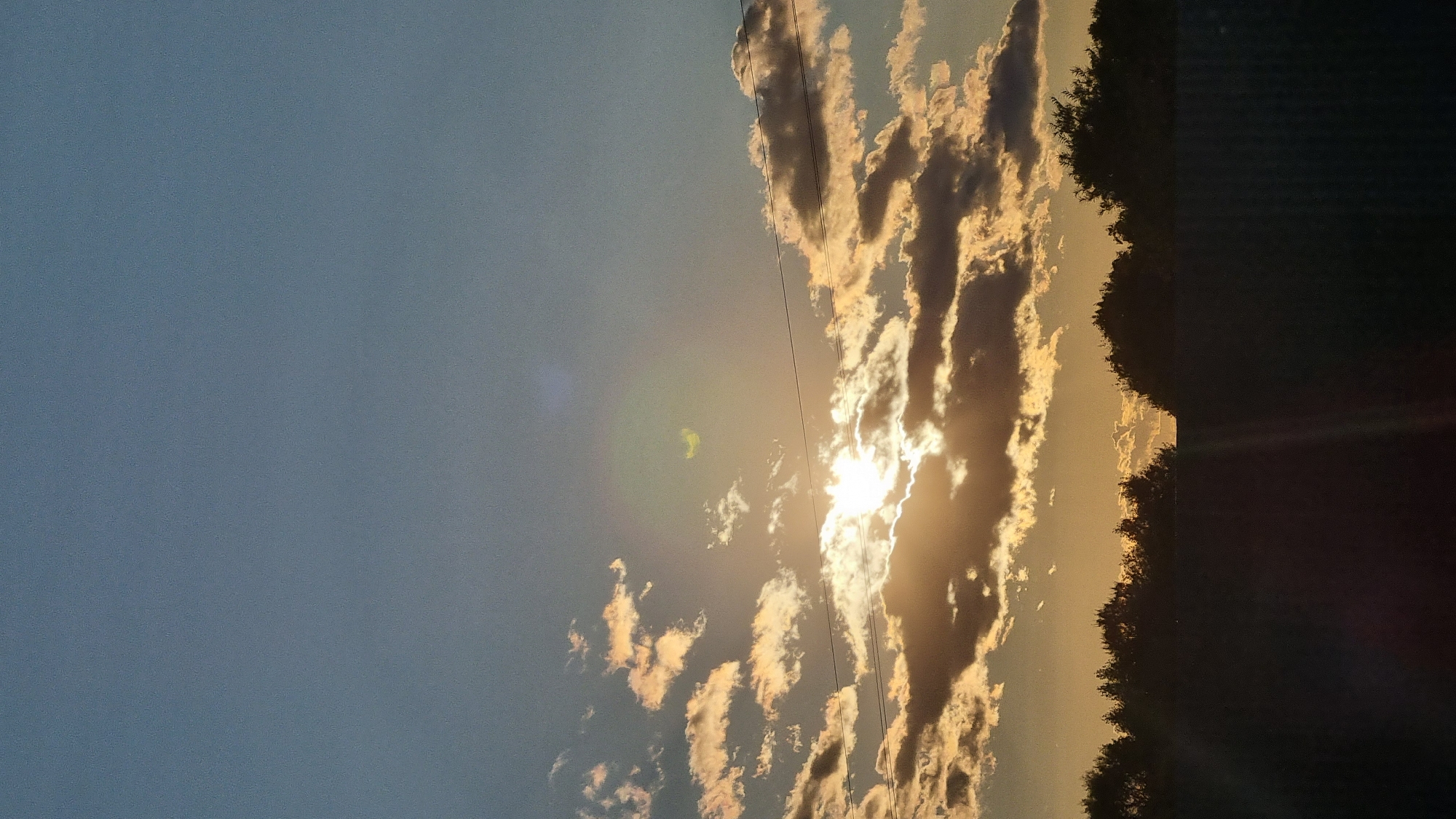 This image shows a sunset over a silhouetted landscape. The sky is a gradient of blue and orange, with the sun partially obscured by clouds. The clouds are illuminated by the sun, creating a dramatic effect. Below the sky, there is a dark silhouette of trees and a fence. There is no visible indication of the location in the city of Vysokoe, Russia.