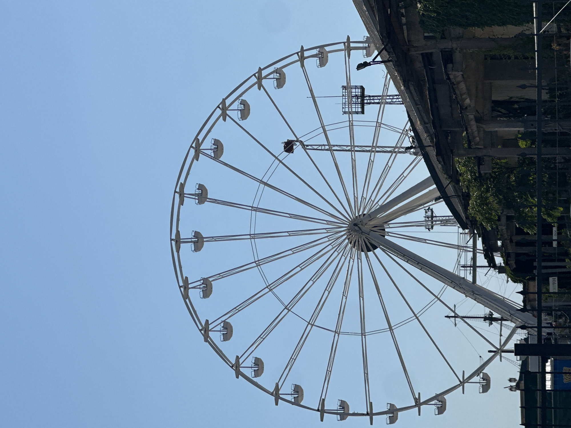 An outdoor, eye-level, medium shot captures a large, white Ferris wheel against a clear, pale blue sky. The Ferris wheel dominates the upper two-thirds of the frame, its circular structure and numerous gondolas clearly visible. The lower portion of the image shows a dilapidated, low-rise building with a damaged roof, exposed concrete supports, and overgrown ivy. A black metal fence runs across the bottom of the frame, obscuring the ground level. Various industrial elements, such as cranes, poles, and what appears to be part of a ship's mast, are visible behind the Ferris wheel and to the right of the building, suggesting a waterfront or industrial area. The overall scene conveys a sense of decay and abandonment, juxtaposed with the grandeur of the Ferris wheel. The lighting suggests it is daytime, likely a sunny day.