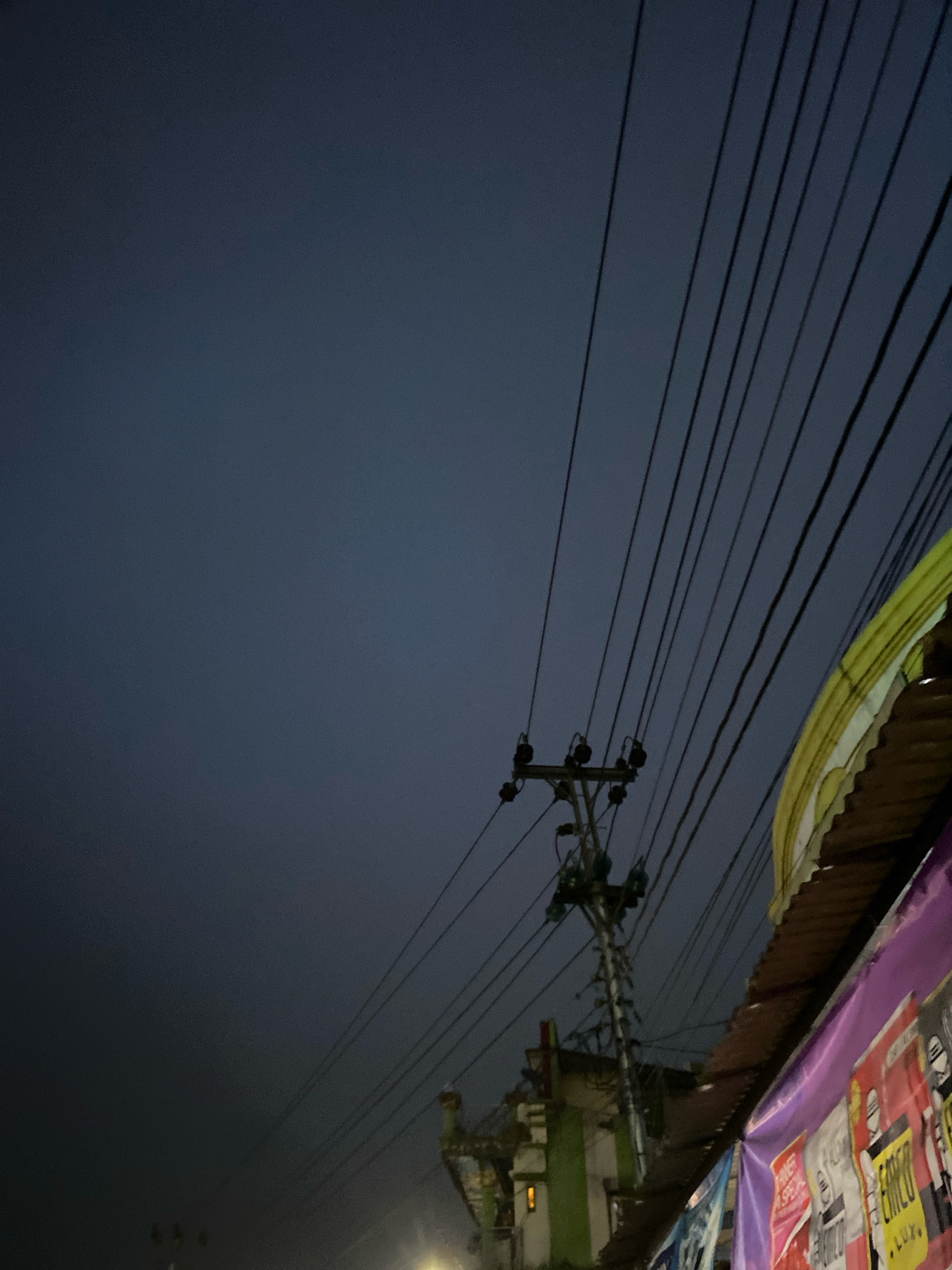 This image depicts a street scene in Kertek, Indonesia, at dusk or night. The sky is a dark, hazy blue, indicating low light conditions. A utility pole with multiple wires is prominently visible, stretching diagonally across the frame. The wires themselves are thin and numerous, creating a sense of visual complexity.
In the lower right portion of the image, the edge of a building or structure is visible. It is covered by a corrugated metal roof, and below that, a purple tarp or banner is adorned with various posters. Some of these posters have text and images, though they are not clearly legible due to the low light and angle. One poster in particular, red and white, appears to be an advertisement or announcement.
A green and white building is partially visible behind the utility pole, with a faint orange light emanating from a window or fixture. The overall impression is one of a typical urban street in Indonesia, likely with shops or residences lining the road. The scene is devoid of people, focusing instead on the infrastructure and the ambient atmosphere.