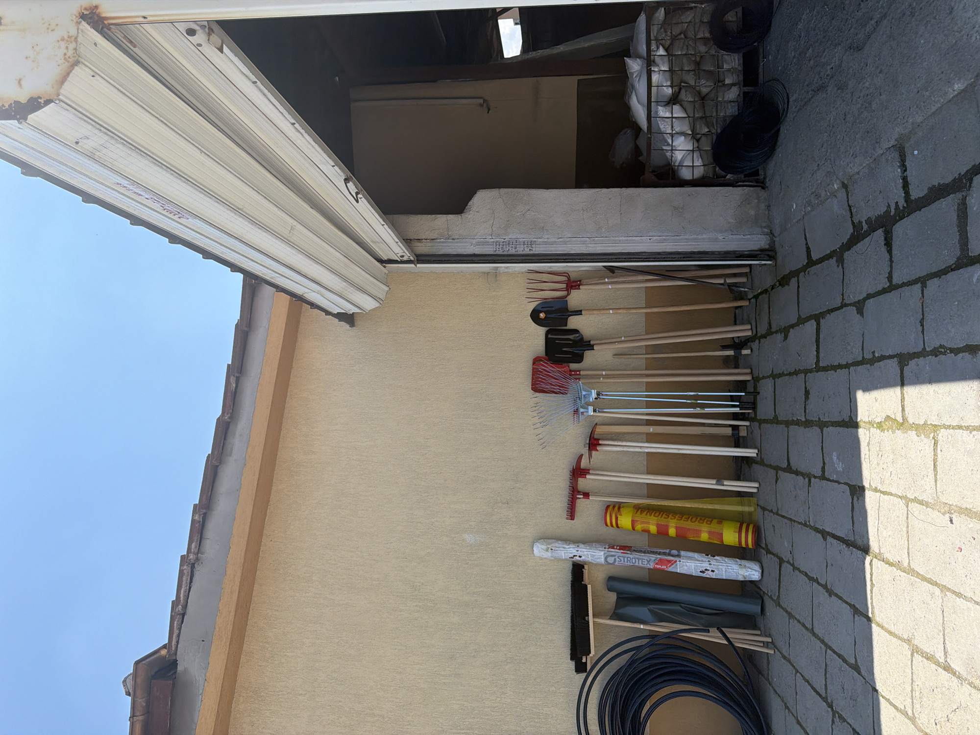 In Zlatitrap, Bulgaria, a collection of gardening tools stands against a tan-colored building under a clear blue sky. To the left, a large coil of black hose is propped up. Adjacent to it are two dark green protective covers and a white roll of material with red text. A yellow and red cylindrical object is next to these. A series of rakes, shovels, and pitchforks with wooden handles are lined up. Some have red metal heads, while others are black and orange. The tools are arranged on a paved surface made of gray interlocking stones. To the right, a white, corrugated metal garage door is partially open, revealing a dark interior with sacks of material piled inside a metal cage. The overall scene depicts an outdoor storage area for tools.