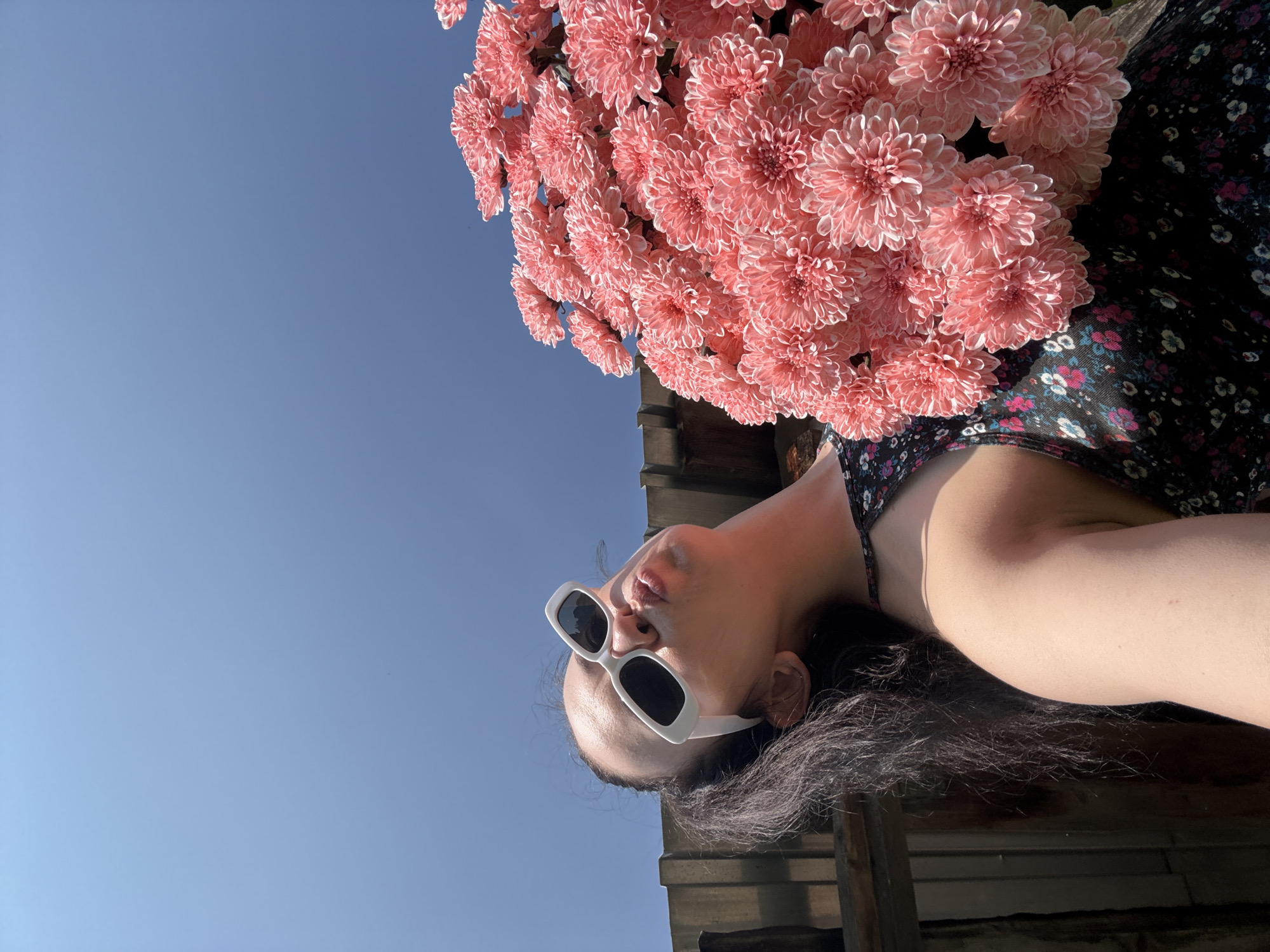An outdoor, low-angle, medium shot captures a woman holding a large bouquet of pink chrysanthemums against a bright blue sky. She is wearing white, rectangular-framed sunglasses and a dark, floral-print dress with a halter neckline. Her lips are puckered, as if blowing a kiss. Her dark hair is pulled back in a ponytail, and some strands fall over her shoulder. The background features a weathered wooden fence and a corrugated metal roof. The image appears to have been taken on a sunny day. The location is identified as Zasechnyi, Russia.