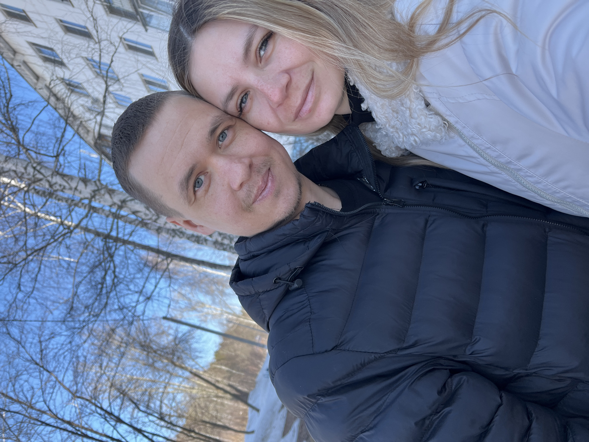 The image displays a close-up, outdoor portrait of two individuals, a male and a female, in the city of Chaikovsky, Russia. The male, positioned on the left, has short dark hair, light-colored eyes, and light facial stubble, wearing a black hooded puffer jacket over a dark long-sleeved shirt. The female, on the right, has long blonde hair, light-colored eyes, and visible freckles across her nose and cheeks, dressed in a light-colored puffer jacket featuring a cream-colored curly fleece collar. Their heads are touching, and both are looking towards the camera with neutral to slightly smiling expressions.
The background consists of a clear blue sky and a sparse stand of bare deciduous trees, including a prominent birch tree trunk visible on the right. Patches of snow are present on the ground in the lower left portion of the frame, indicating cold weather conditions. In the upper right background, a multi-story white residential building with numerous windows is partially visible, indicating a developed urban or suburban environment within Chaikovsky. The scene is consistent with a late winter or early spring setting in Russia.