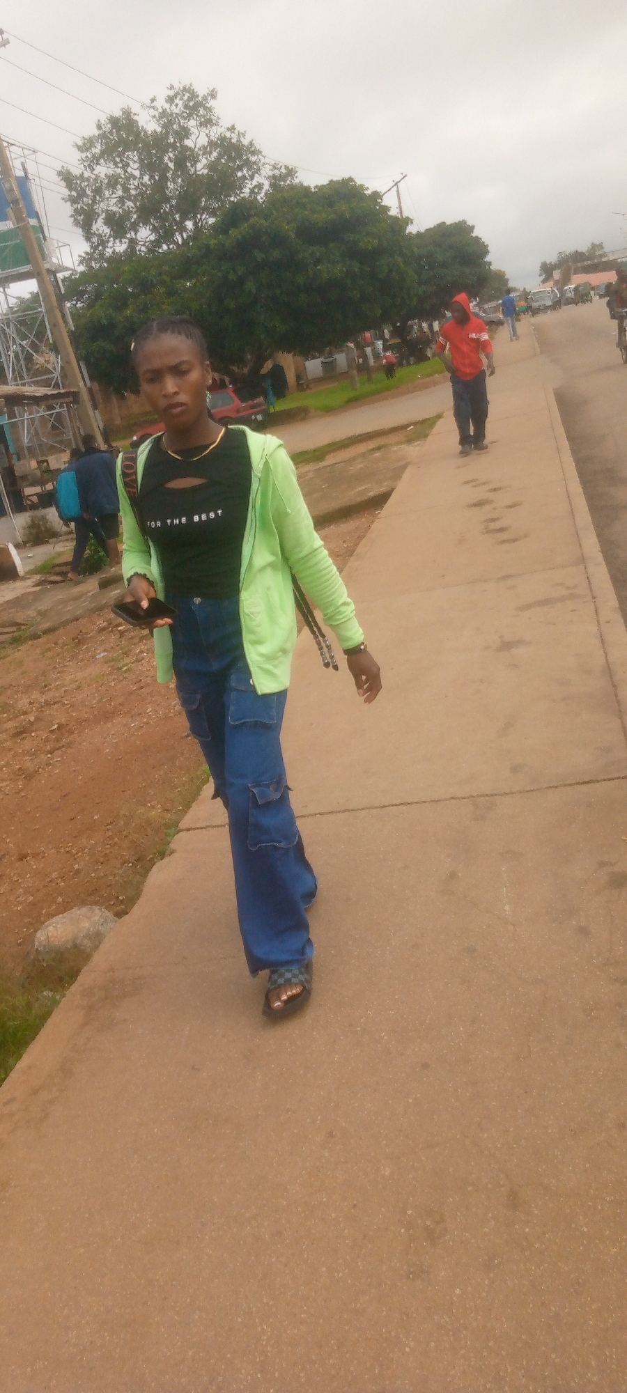 A young woman is walking on a sidewalk in an outdoor urban environment. She is wearing a black crop top with the words "FOR THE BEST" written in white, a light green zip-up hoodie, and blue baggy cargo pants. She is also wearing checkered slide sandals and has a black smartphone in her left hand. Her hair is pulled back in braids. In the background, there are trees, a few other people, some vehicles, and buildings. The sky is overcast.