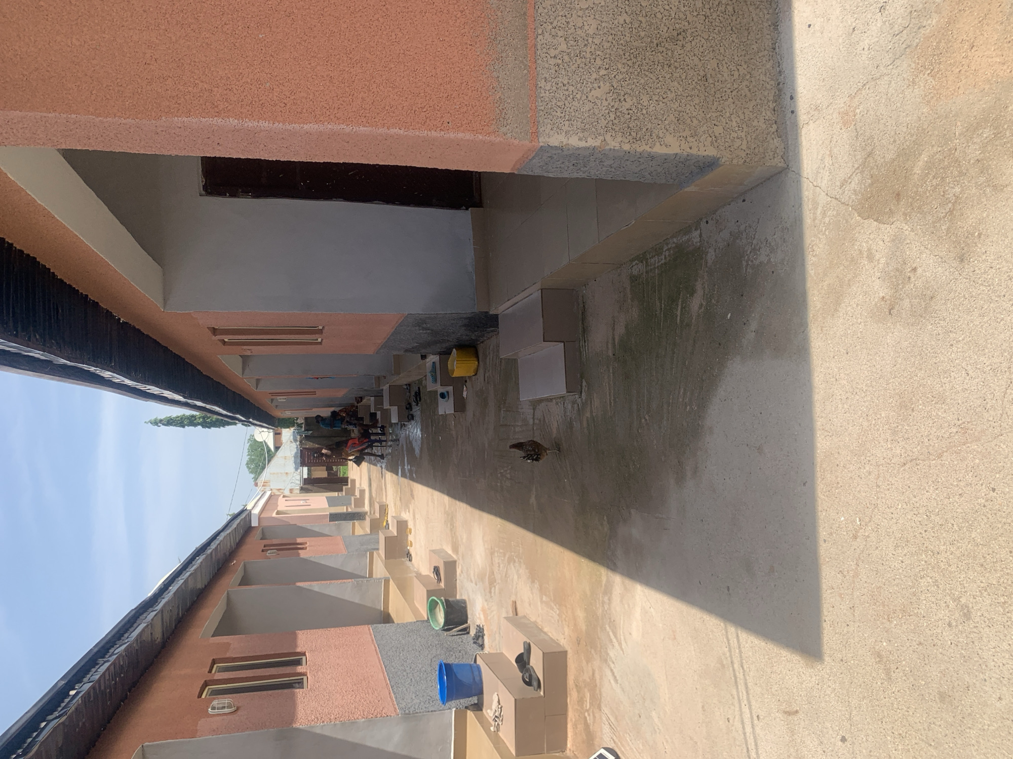 In Jauro Yinu, Nigeria, a narrow walkway separates two buildings with orange and grey textured walls and corrugated metal roofs. The walkway is paved with concrete and is partially wet, suggesting recent activity or rain. On the left side, several steps lead up to the entrance of a building. A blue bucket and a green bucket are placed on the steps, along with a pair of black sandals and a folded piece of cloth. Further down the walkway, a chicken stands on the wet concrete. On the right side of the walkway, more steps lead to doorways. In the distance, several people are gathered, some appear to be sitting on the steps. A tall, slender tree is visible in the background, and the sky is clear and bright blue. The scene appears to be an outdoor residential or commercial area.