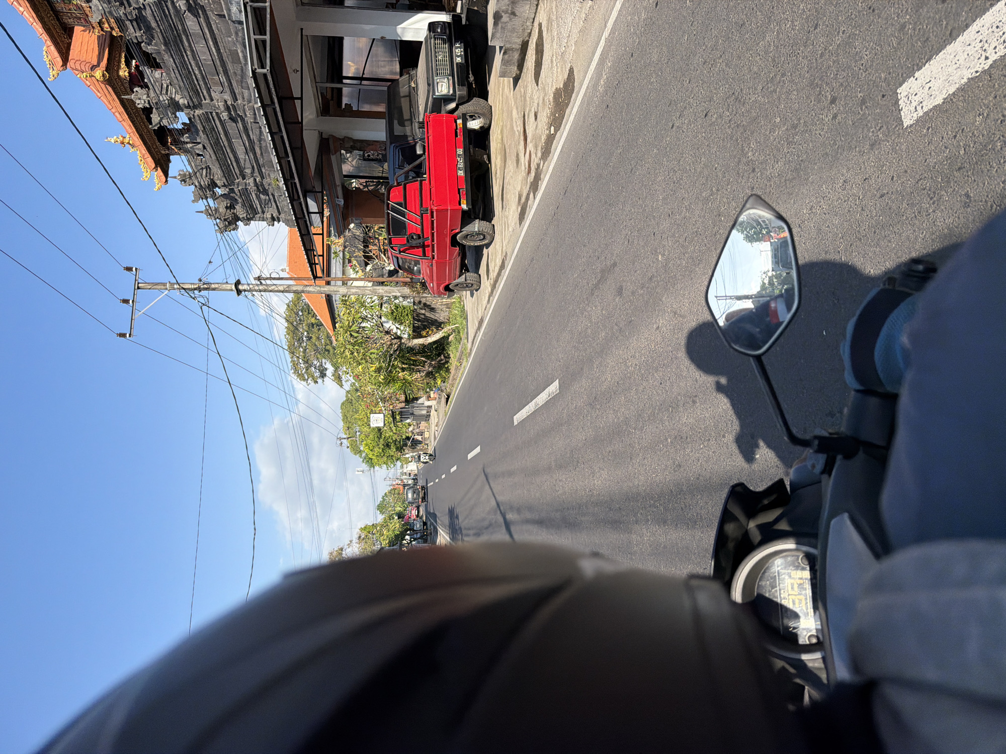 A first-person perspective shot from a motorcycle in Mengwi, Indonesia shows a street scene on a clear day. In the foreground, a portion of the motorcycle rider's helmet and shoulder is visible, along with the motorcycle's handlebars and instrument panel. The road ahead is paved with asphalt and marked with white lines. On the right side of the road, a traditional Balinese building with intricate stonework and an orange tiled roof stands prominently. Next to it, parked on the sidewalk, are a red pickup truck and a dark-colored SUV. The red truck has a license plate that reads "DK 8245 DD." Further down the road, other vehicles and buildings can be seen, as well as trees and power lines crisscrossing the sky. A motorcycle rider is visible in the distance on the right side of the road, and another is reflected in the motorcycle's side mirror. The shadow of the rider and motorcycle is cast on the road.