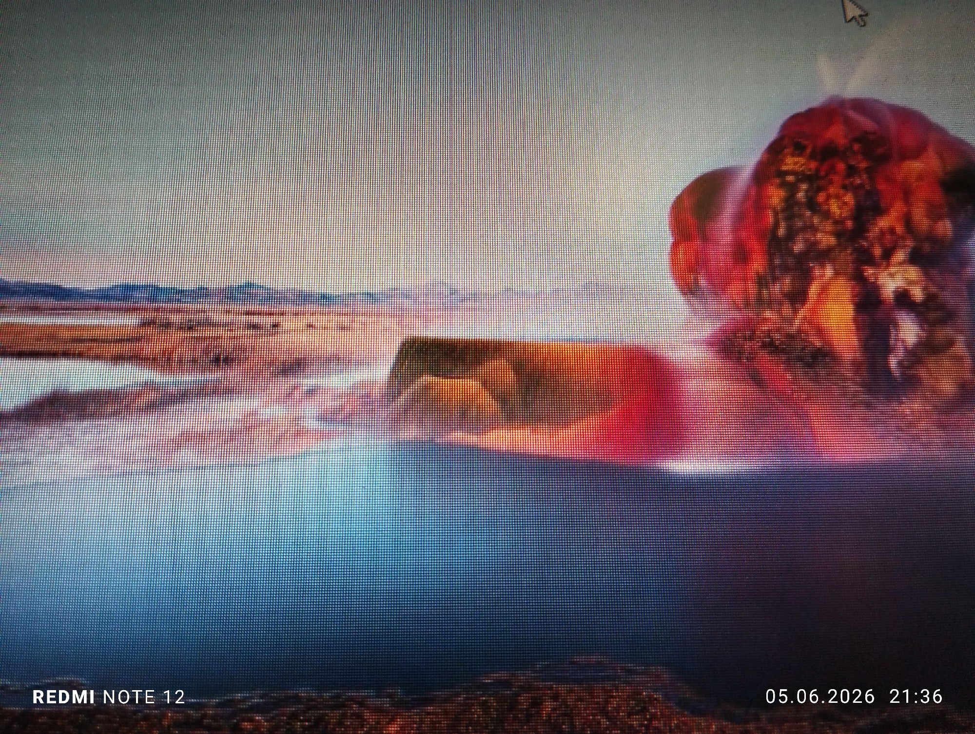 A photograph captures a digital screen displaying a highly pixelated, abstract landscape. The screen exhibits a distinct grid pattern characteristic of a display. The upper portion of the screen depicts a pale, hazy sky transitioning into an indistinct horizon line with dark, muted shapes suggestive of distant landmasses. Below the horizon, a wide expanse of warm, earthy tones (oranges, reds, browns) is visible, with a central horizontal band of darker, solid forms. The lower half of the screen is dominated by a deep, dark blue, suggesting water. On the right side, a large, vibrant, irregular formation, predominantly red and orange with darker, textured internal areas, dominates the mid-to-upper right, evoking an explosion or dramatic natural phenomenon. A white computer mouse cursor, shaped as an arrow, is visible in the upper right quadrant, pointing slightly upwards. Text in the lower left corner reads "REDMI NOTE 12", indicating the capture device. Text in the lower right corner reads "05.06.2026 21:36", indicating the date and time of capture. The image was captured in the city of Burgas, Bulgaria.