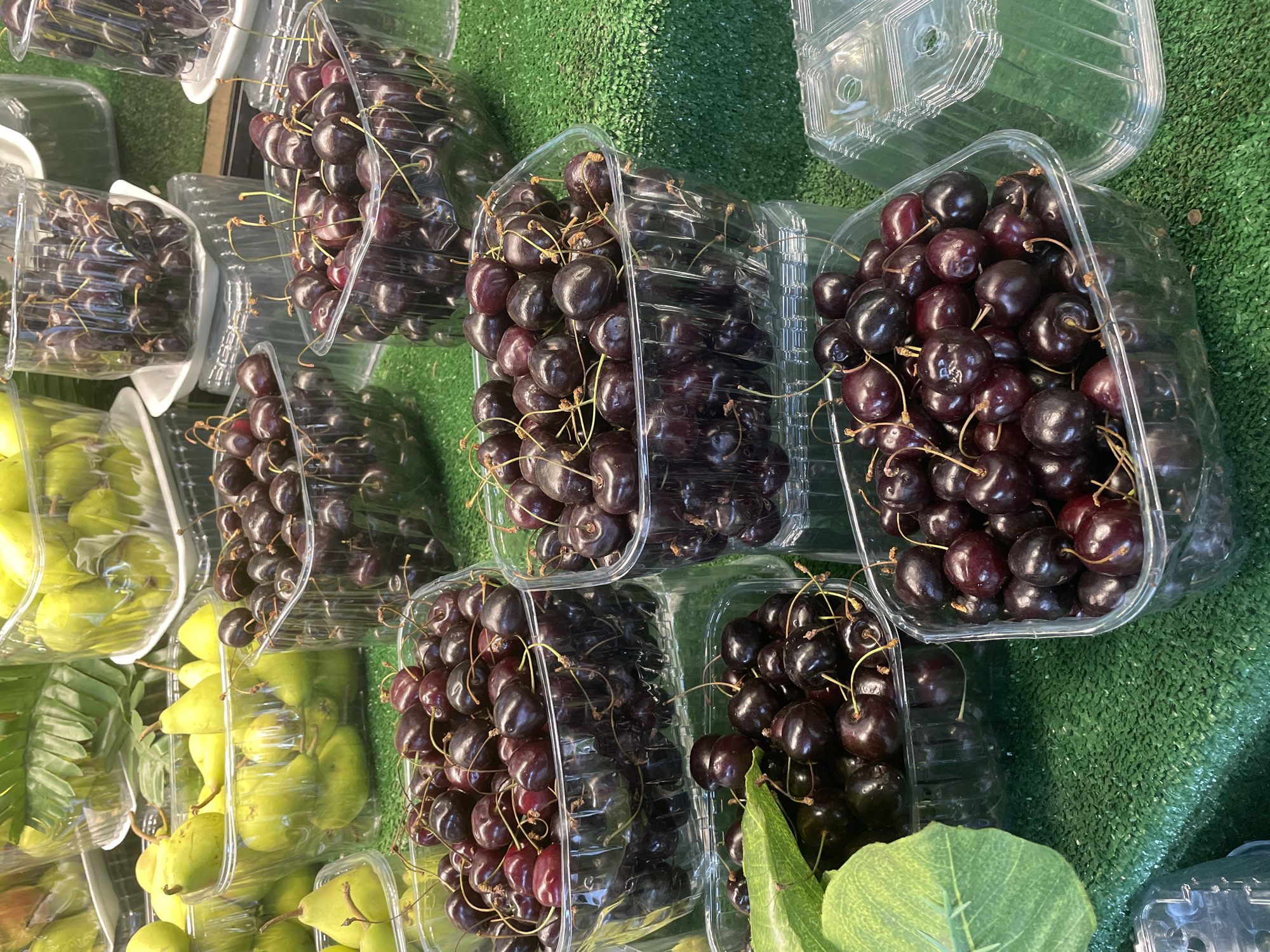 In Kadıköy, Turkey, a market stall displays an assortment of fresh fruit. The foreground features several clear plastic containers overflowing with dark red cherries, each with slender stems. Behind the cherries, a variety of light green pears are visible in similar plastic packaging. The fruit is arranged on a green, artificial turf-like surface, suggesting a market or produce stand environment. The lighting is bright and even, highlighting the vibrant colors and textures of the fruit.