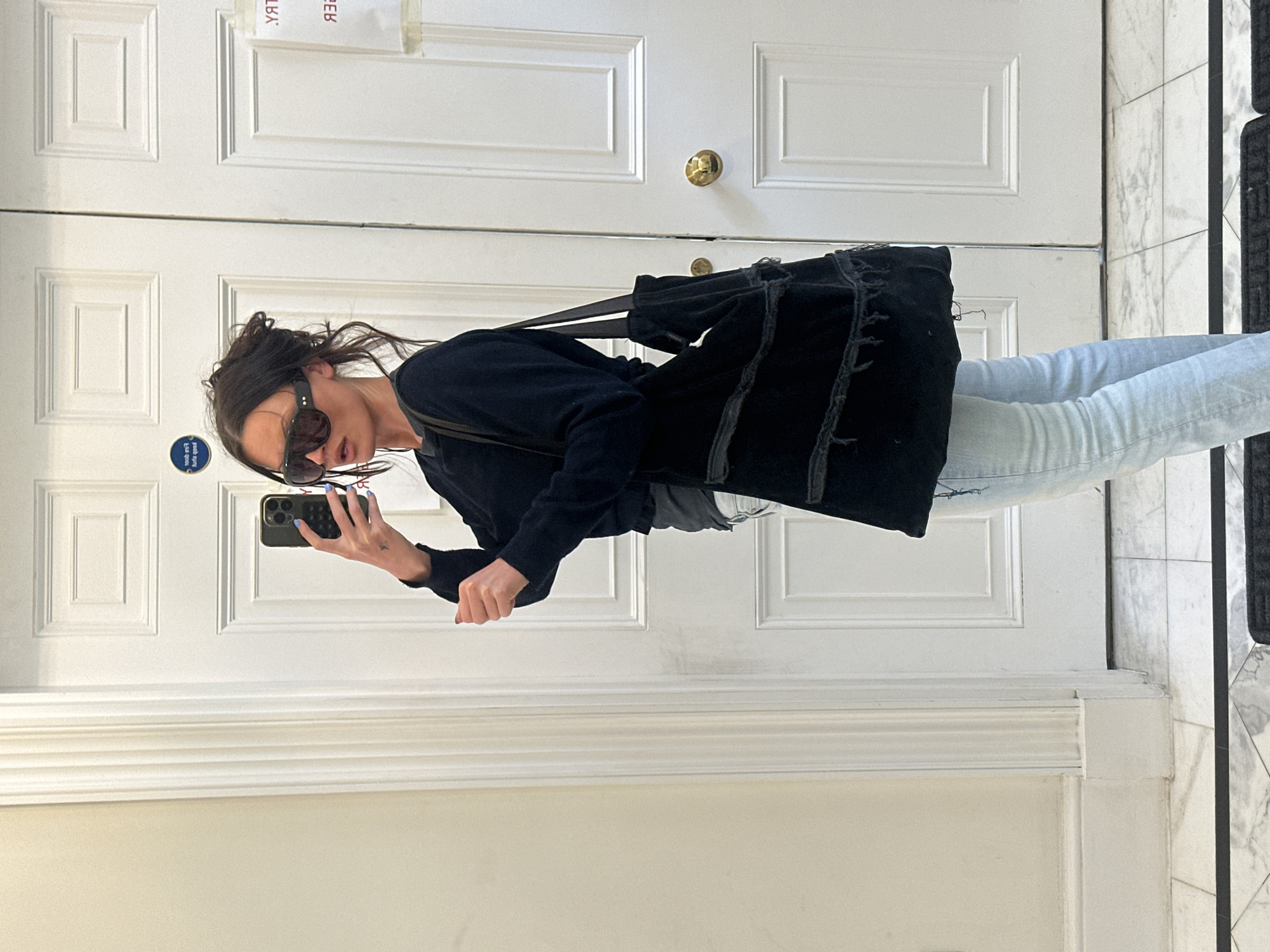 The image captures a mirror selfie of a person standing indoors. The subject, seen from the waist up, is wearing a dark long-sleeved top and light blue denim jeans. They have dark hair styled in a messy updo and are wearing dark sunglasses. A black tote bag with a distressed, frayed fringe design hangs from their shoulder. The person is holding an iPhone (identifiable by its distinct camera array) in their right hand, with light blue nail polish visible on their fingernails.
The background, which is a reflection in the mirror, shows a white paneled door with a brass doorknob. Below the door, white wainscoting with decorative moldings lines the lower part of the wall. To the right, a patterned marble floor in shades of grey and white is visible, alongside a segment of a black doormat.
Several pieces of reversed text are visible on the door's reflection. On the upper left, a partially obscured label shows "YЯAT" and "ƎB". Below this, a small circular blue sticker with white reversed text is also present, though its specific content is illegible. The setting appears to be an interior hallway or entryway of a building, likely during daytime given the general brightness. This type of interior is common in residential or commercial buildings in London, United Kingdom.