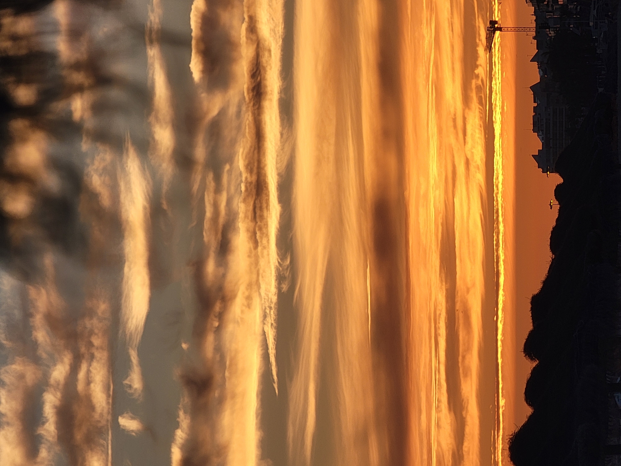 The image depicts a dramatic sunset over Quarteira, Portugal. The sky is filled with layers of colorful clouds, ranging from deep orange and yellow near the horizon to darker grays and browns higher up. The clouds are streaked and wispy, indicating movement and varying altitudes.
In the foreground, the dark silhouettes of beach umbrellas and lush vegetation create a textured, undulating line across the bottom of the frame. Beyond this, a city skyline is visible in silhouette against the bright sky. Several buildings of varying heights and architectural styles can be discerned. A prominent construction crane stands to the right of the frame, its skeletal structure stark against the vibrant sky. The overall impression is one of a serene yet dynamic end to the day.