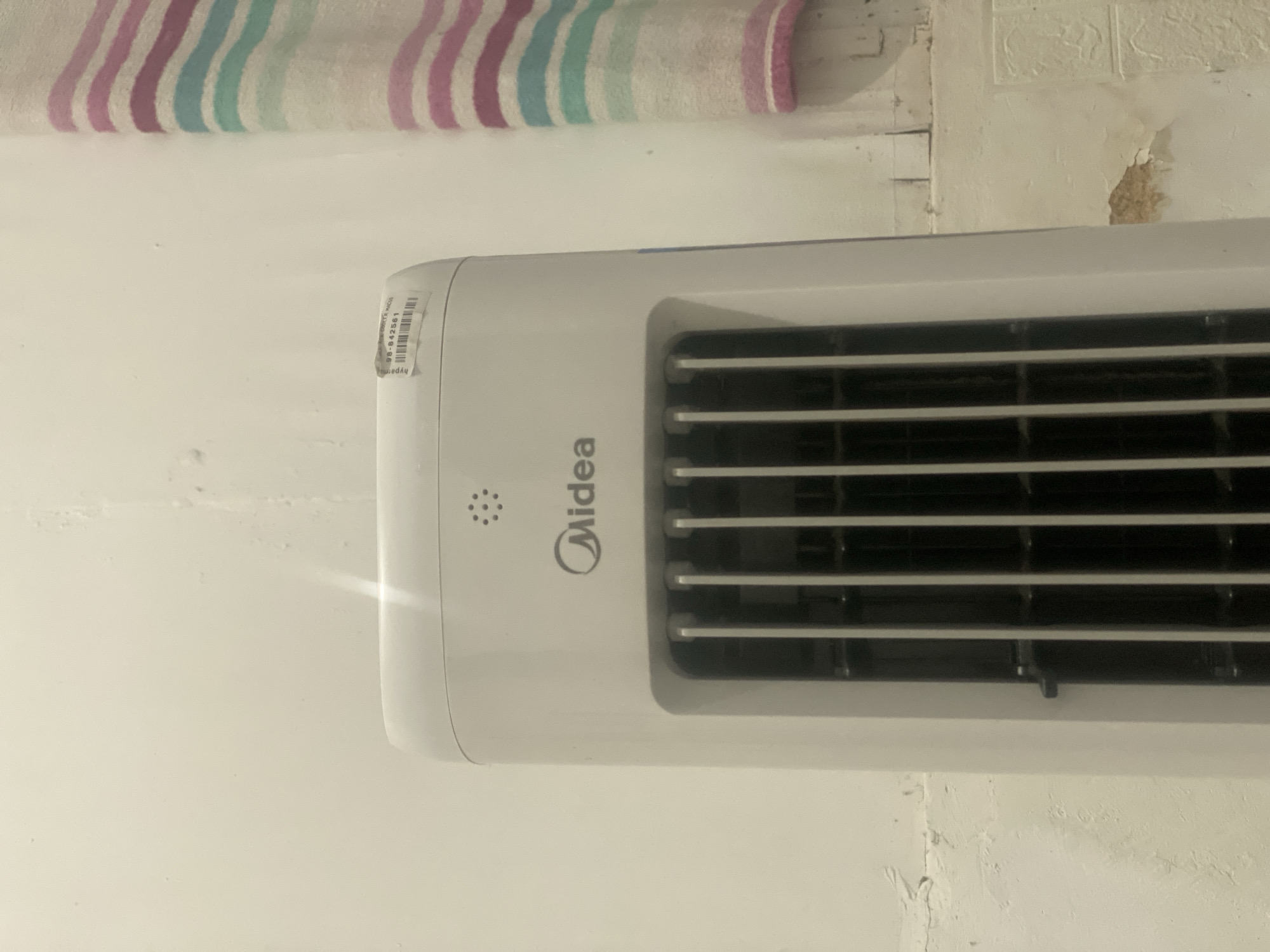 The image shows the top portion of a white Midea brand tower fan, placed indoors against a wall. The fan has a vertical grille and a small circular vent above the Midea logo. A sticker with a barcode and the numbers "98-842561" is affixed to the top right of the fan. To the right of the fan, a vertically striped fabric, possibly a towel or curtain, with bands of pink, blue, and white, hangs against the wall. The wall itself appears to be a light color with some textured patterns and a peeling section on the lower right. The lighting suggests an indoor environment. The location is Banjarbaru, Indonesia.