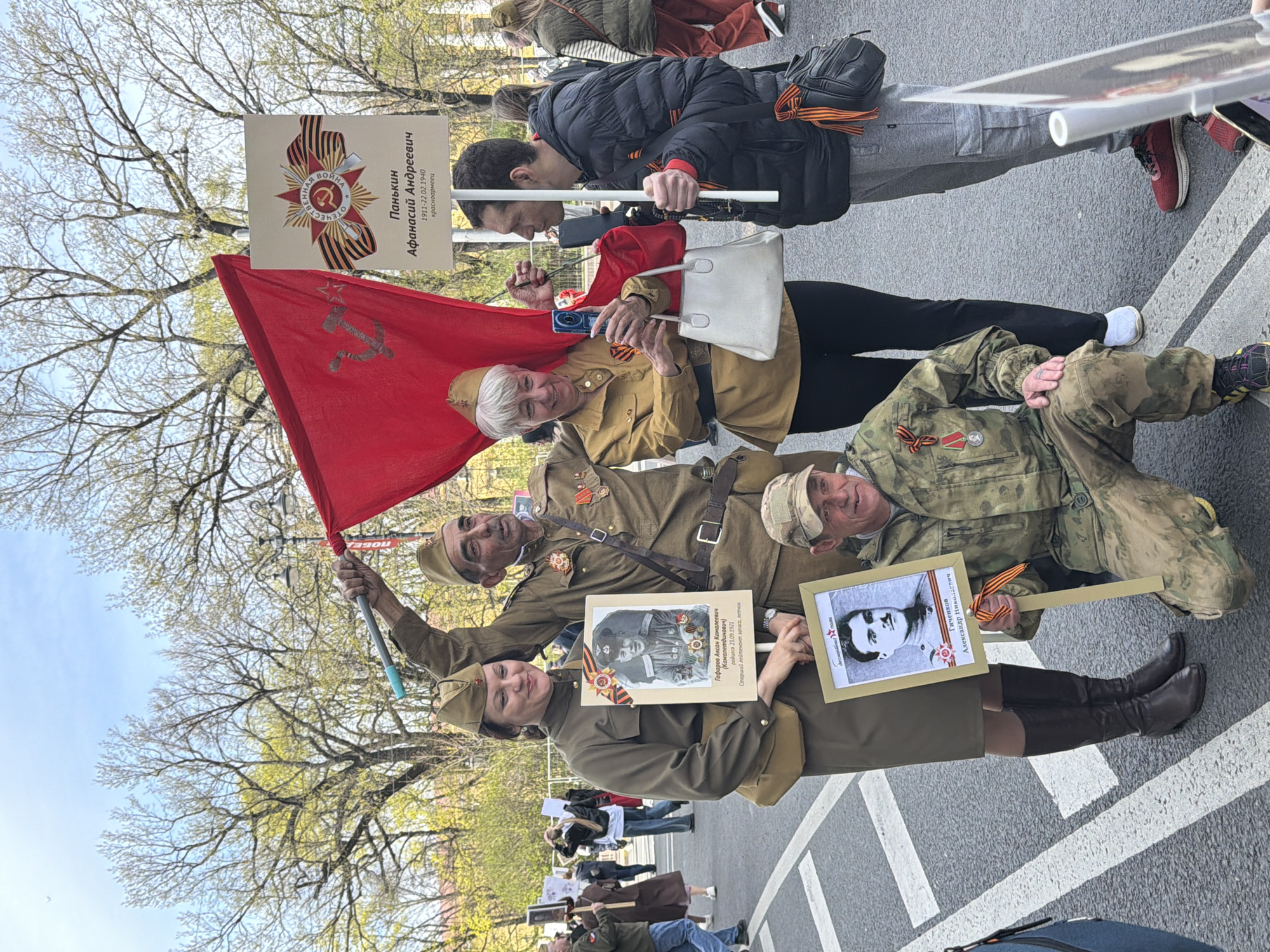 The image captures a group of people participating in a parade or march, likely commemorating Victory Day on May 9, a Russian holiday that celebrates the Soviet Union's victory over Nazi Germany in World War II. The location appears to be in a city, possibly St. Petersburg, Russia.
Five people are prominently visible in the foreground of the image, walking in a row. They are holding portraits of deceased soldiers and a red flag bearing the hammer and sickle symbol. They are dressed in Soviet military uniforms from the World War II era, including hats, tunics, belts, and boots. Some of them also have medals pinned to their uniforms.
One person is holding a red flag with the communist hammer and sickle. The other people hold signs that have photos of soldiers with writing in Russian. The signs identify the name, ranks, and birth dates of the soldiers.
The event appears to be taking place on a paved road or street. There are other people participating in the event in the background. The sky is overcast, and the trees are in bloom.