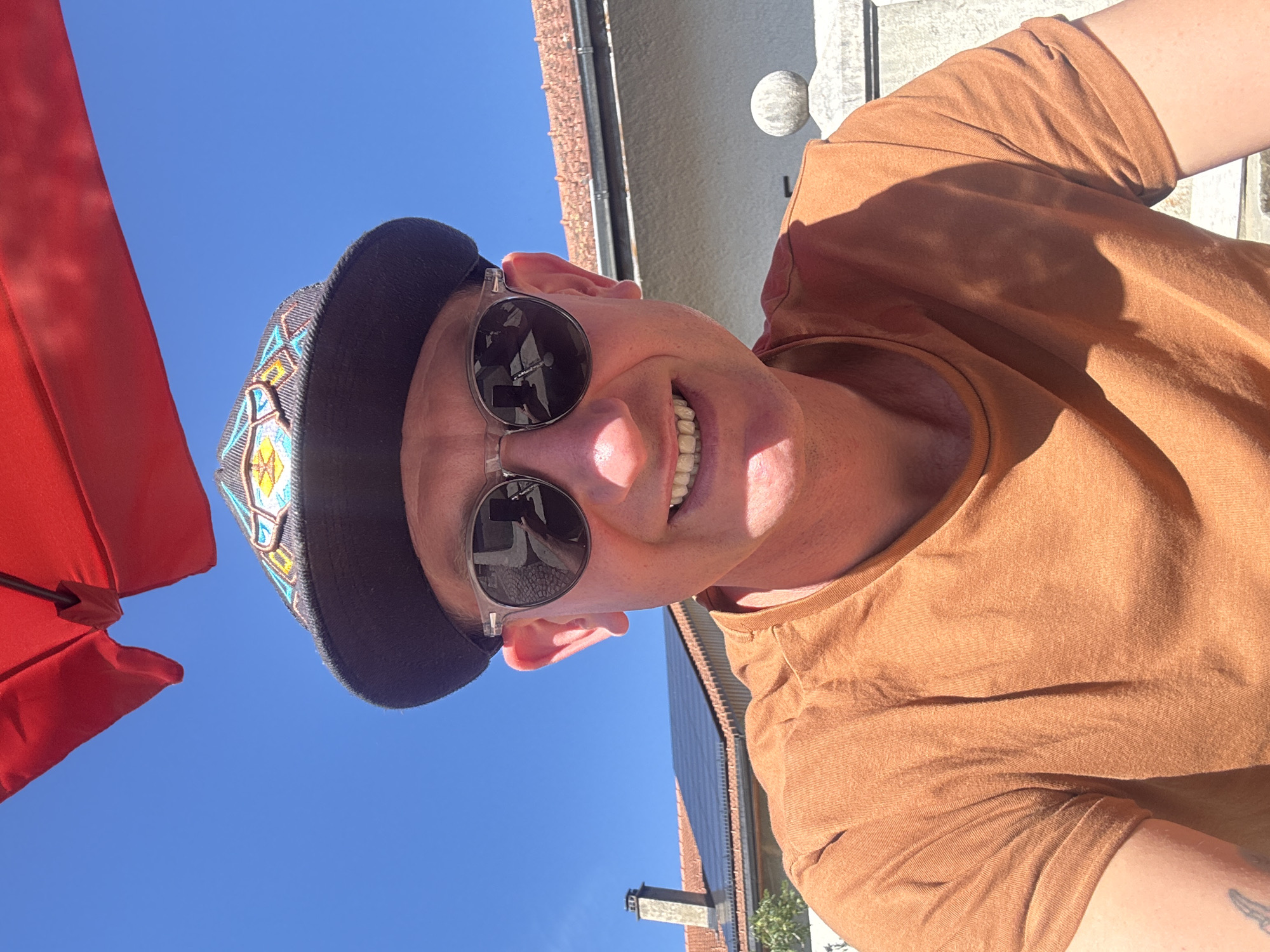 A man wearing a black baseball cap with a colorful geometric pattern, clear-rimmed sunglasses, and an orange t-shirt smiles at the camera. He is outdoors, with a bright blue sky above and the partial view of a red umbrella. In the background, there is a house with a red tile roof and solar panels, and a stone pillar. The scene appears to be in Petit-Val, Switzerland.