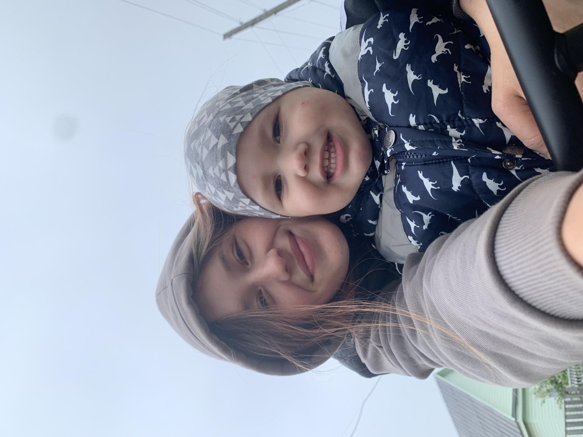 This close-up, upward-facing selfie captures an adult and a young child outdoors. The adult, likely female, has long brown hair and wears a grey hooded top, smiling gently at the camera. The child, a toddler or baby, is nestled in front of the adult, wearing a grey beanie with a geometric pattern and a navy blue puffer jacket adorned with white dinosaur silhouettes. The child has a wide, joyful smile, revealing small teeth, and shows a few small red spots on their cheek and forehead.
Both subjects are looking directly at the camera, conveying happiness and affection. The background consists of a bright, overcast sky, suggesting daytime with diffused light. Faint structures like utility wires or poles and a part of a light green building or roof are vaguely visible in the distance. The child's jacket features buttons with the faint text "ORIGINS" on them. The cool-weather attire (puffer jacket, beanie, hooded top) indicates a cooler climate. The photo captures a tender, happy moment between the two in Фоки, Russia.