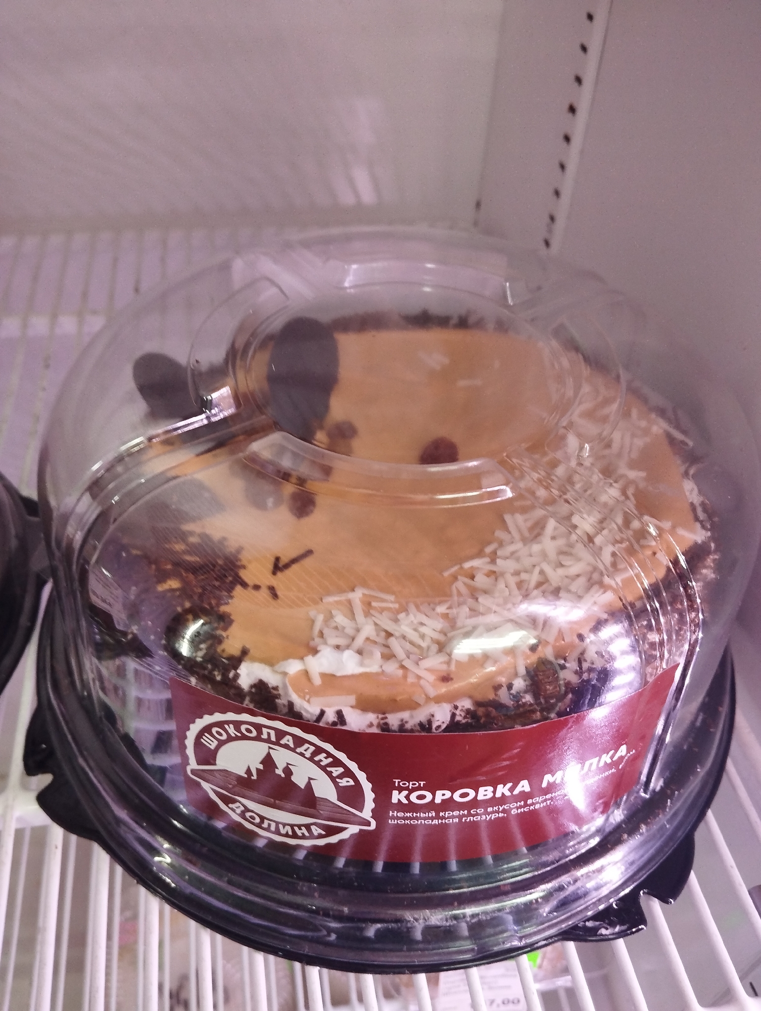 A packaged cake sits on a white, wire shelf, likely inside a refrigerator or display case. The cake has a caramel-colored top with small, dark brown decorations and sprinkled with white, shredded coconut. A white cream frosting is visible along the edge. The cake is packaged in a clear plastic dome on a black plastic base. The label on the cake has a maroon background and features the name "Шоколадная Долина" in Russian, along with a picture of a castle or landmark. The cake's name is "КОРОВКА МОЛОКА," also in Russian. The label also mentions "Торт," indicating this is a cake. The writing on the label suggests a "delicate cream with the taste of boiled chocolate glaze, biscuit." The shelf is likely inside a store or market, and is in Krasnogorskoye, Russia. The price tag, "7.00", is visible at the bottom.