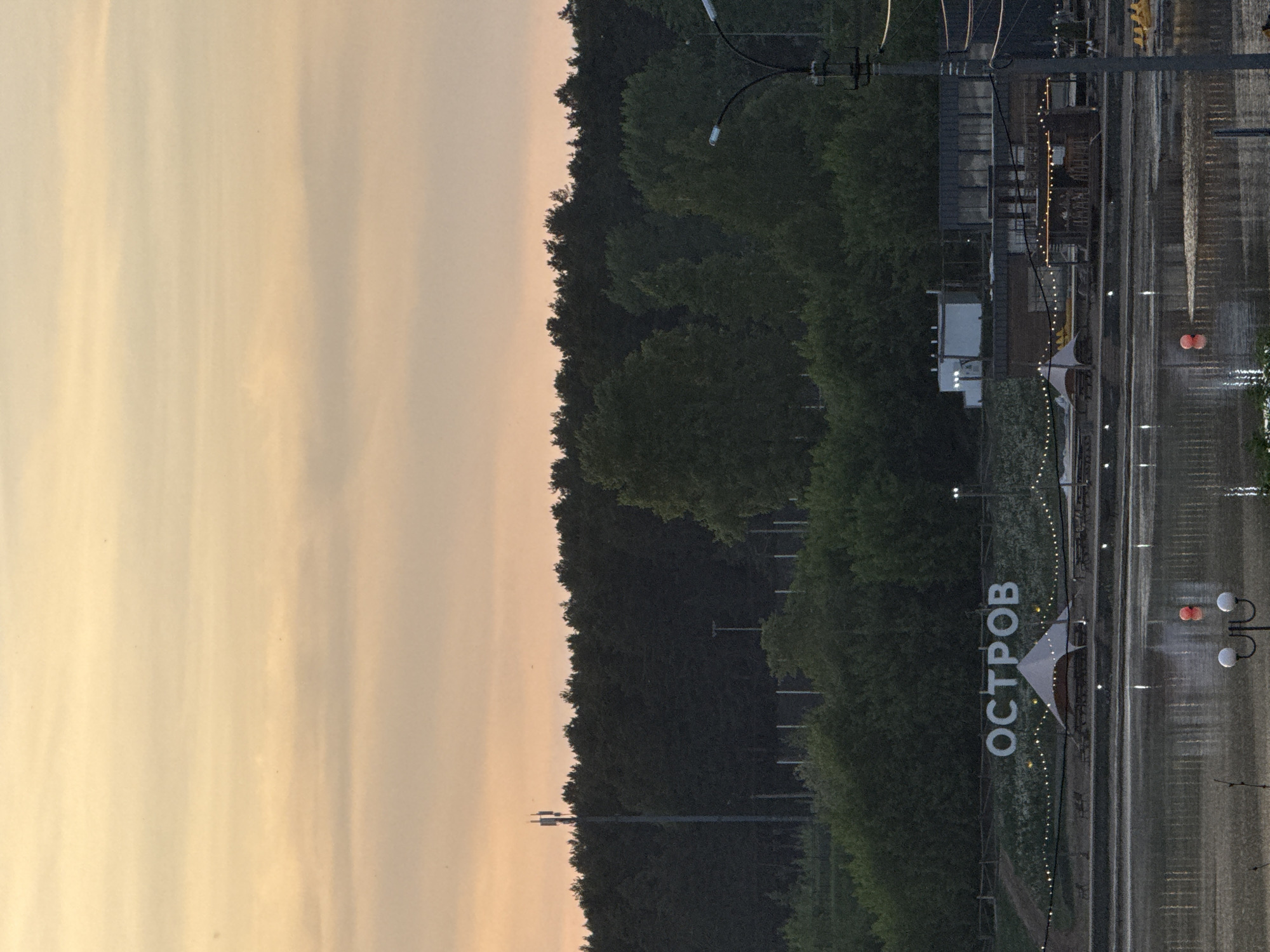 The image shows a view in Votkinsk, Russia. On the right side, there is a body of water. To the left of the water is a sign that says "ОСТРОВ" (OSTROV), which means "Island." There is a small white tent structure near the sign. Lights are strung up overhead. Trees are visible along the middle of the image, separating the sky on the left from the waterfront on the right. The sky has a yellow-orange hue suggesting either sunrise or sunset. Power lines are visible above the trees and the waterfront area. There's a building or structure further back from the water, also with lights strung around it. Street lights are visible as well.