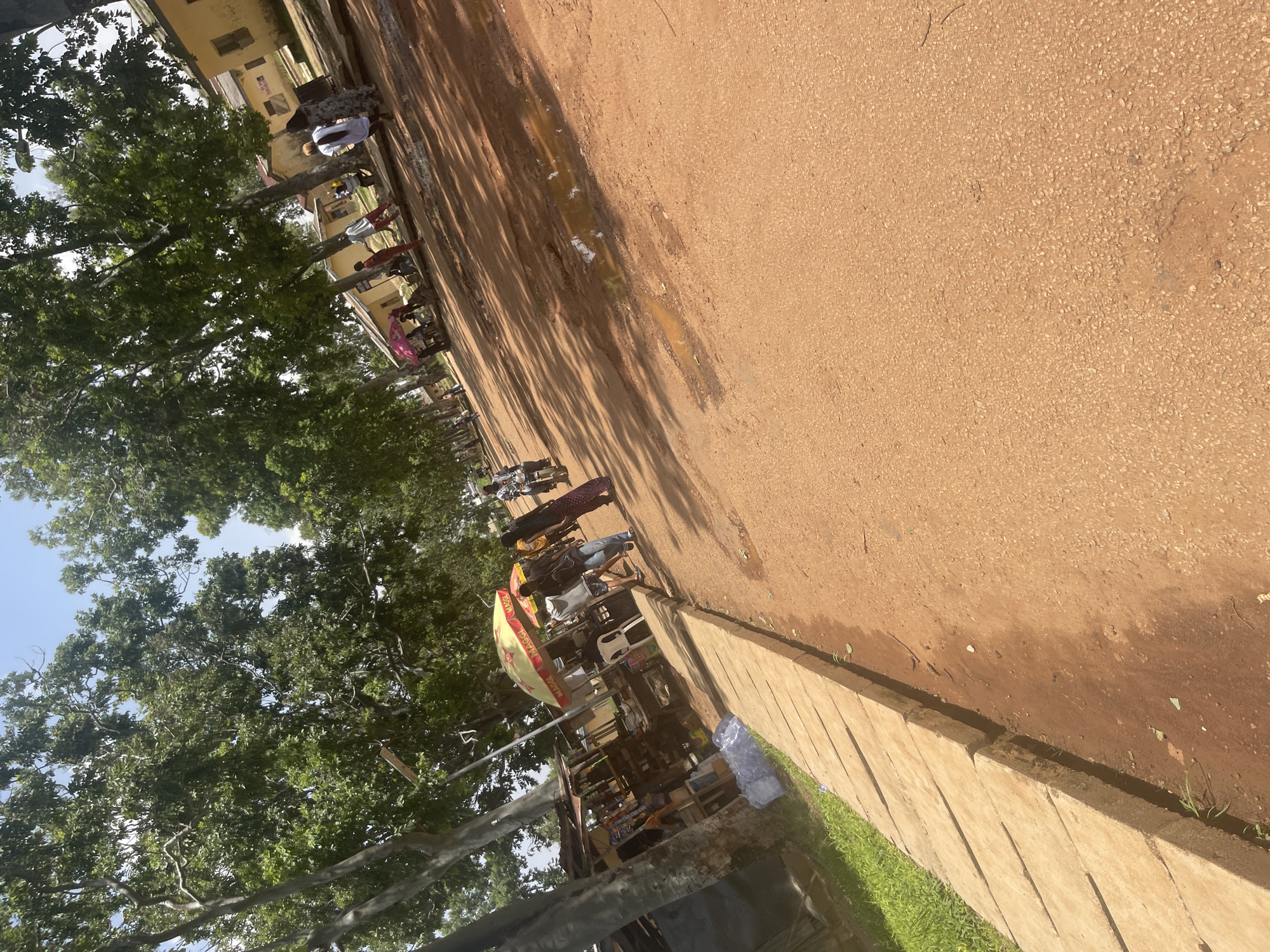 The image depicts a bustling street scene in Rafin Par, Nigeria. A dirt road, characterized by its reddish-brown hue and scattered puddles, dominates the foreground and stretches into the distance. On the left side of the road, a concrete sidewalk runs parallel to a row of small shops and stalls, some covered by corrugated metal roofs. Bags of what appear to be water sachets are stacked near one of the stalls. Tall, leafy trees line the street, casting dappled shadows on the ground.
Several people are visible on the street. A group of individuals walks along the sidewalk, some carrying bags. Further down the road, more people are gathered around stalls and in front of buildings. One person is seated under a yellow umbrella with "MAGGI" branding. Another person is seen riding a motorcycle. Buildings with yellow walls and window frames are visible in the background. The sky above is clear and blue. The overall impression is one of a lively, everyday street environment with commercial activity and pedestrian and vehicular traffic.