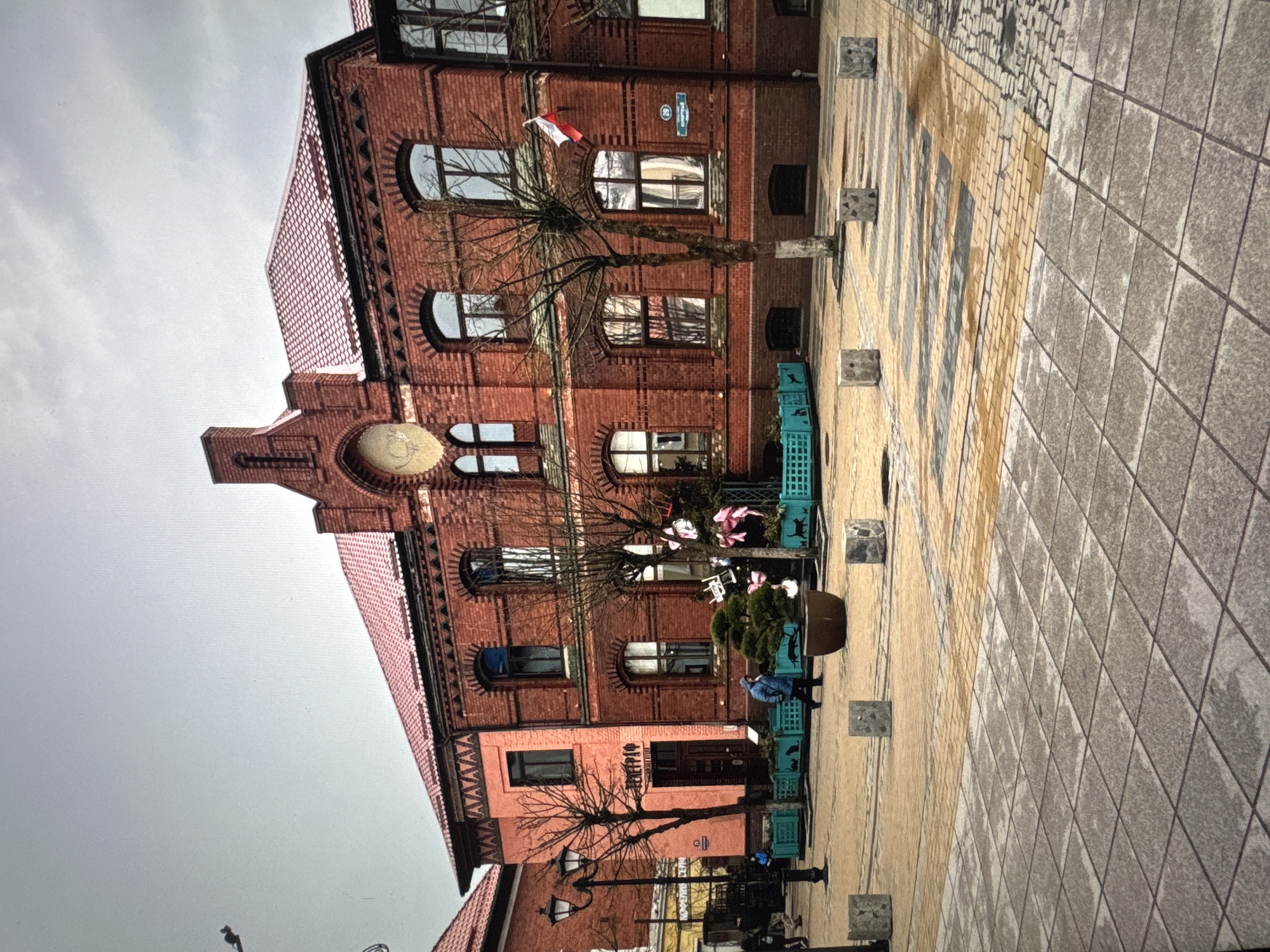 The image depicts a multi-story brick building in what appears to be a town square or pedestrian area. The building has a clock face on its facade and arched windows, suggesting historical architecture. A Polish flag is visible. Trees, benches, and decorative planters line the walkway in front of the building. Several people are present. The pavement is a mix of brick and tile. The sky is overcast. The presence of the Polish flag and architecture are clues. A storefront at the bottom of the brick building features a text sign above the entrance, but the language and content are difficult to decipher. While the request specifies the location as "Новинки, Russia", this is unlikely to be correct, because this area and style of architecture does not match any part of russia.