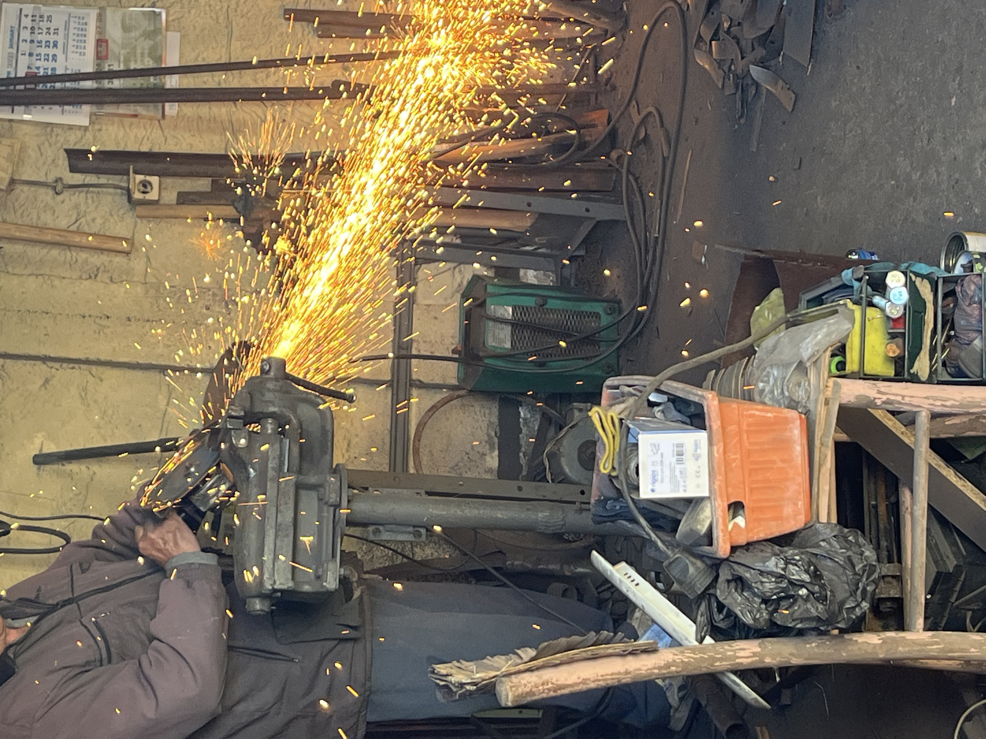 A man is grinding metal in a workshop in Unknown, Unknown. He is wearing a gray jacket and is holding a grinding tool against a metal object held in a vise. Sparks are flying from the grinding point. The workshop appears cluttered, with various tools, metal pieces, and equipment scattered around. A calendar hangs on the wall in the upper left corner.