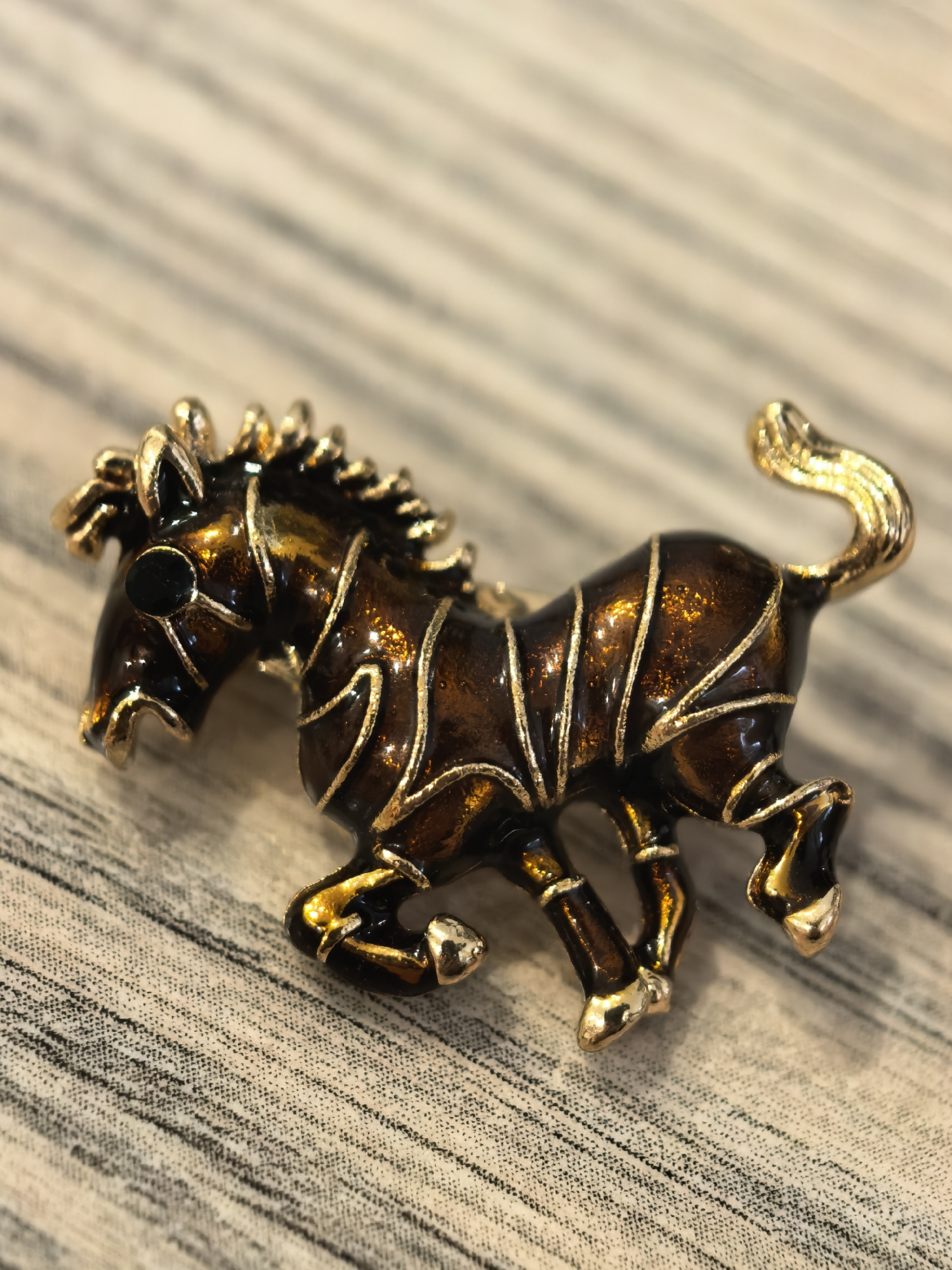 A detailed close-up shot showcases a gold and dark brown enamel brooch shaped like a zebra. The zebra is depicted in profile, mid-stride, with its head lowered and mane flowing. The brooch features intricate detailing on its body, with thin gold lines accentuating the zebra's stripes. The object rests on a textured surface, possibly a fabric or wood grain, with a muted background. There is no indication of the city of Sterlitamak, Russia, or any people in the image.