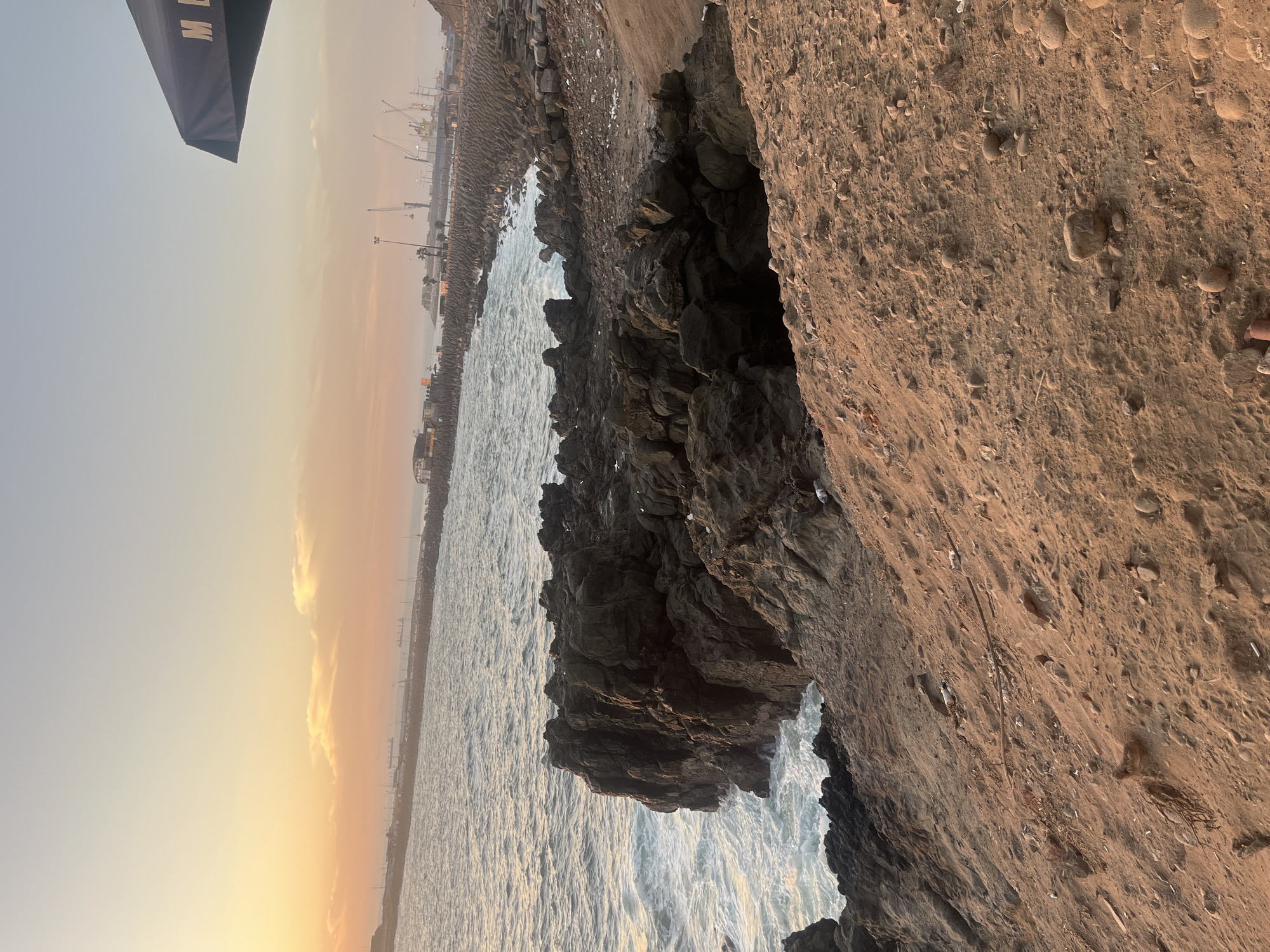 The image depicts a rocky coastline in Arica, Chile, likely at sunset. The foreground features jagged, dark rocks being battered by waves, with a sandy cliff face to the right. In the distance, a pier or port facility with cranes is visible along the horizon. The sky is gradient, transitioning from a light blue to a warm orange near the water. The image is rotated 90 degrees counterclockwise.