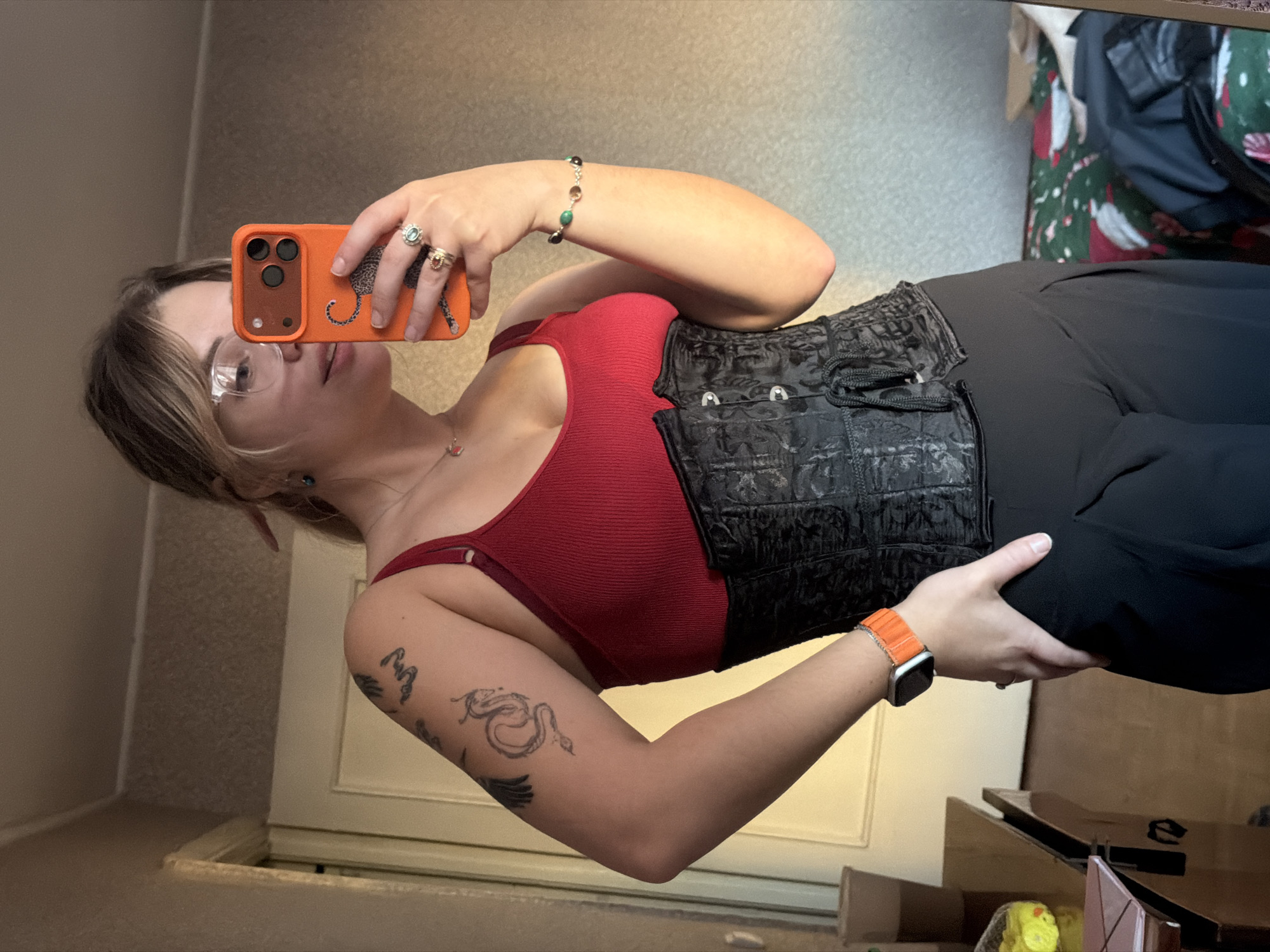 A woman is taking a selfie in a room. She's wearing glasses, a red tank top, a black corset, and black pants. She has an orange phone case with a leopard design. She's wearing several rings and a beaded bracelet. Her left arm has a snake and other tattoos. She's also wearing an Apple watch with an orange band. The background includes a mirror, a door, and what appears to be a cluttered corner with boxes and other items. It is impossible to determine the location of Naberezhnye Chelny, Russia from this image.