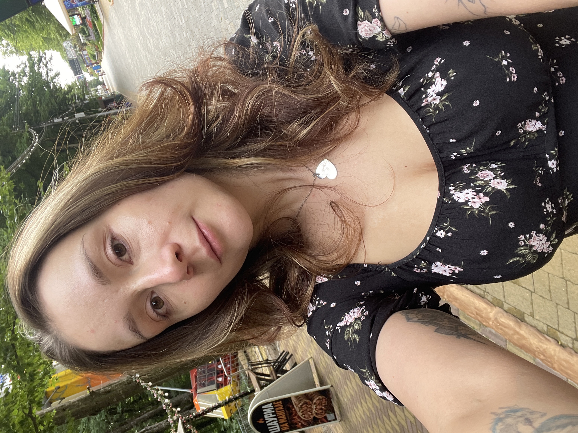 A young woman with medium-length brown hair is taking a selfie outdoors. She is wearing a black dress with a floral pattern and a silver heart-shaped necklace. She has tattoos on her left arm. In the background, there is a paved walkway, trees, and some amusement park rides or stalls. A sign with Cyrillic text is visible on the left. The scene appears to be a public outdoor area, possibly a park or recreational zone in Stavropol, Russia.