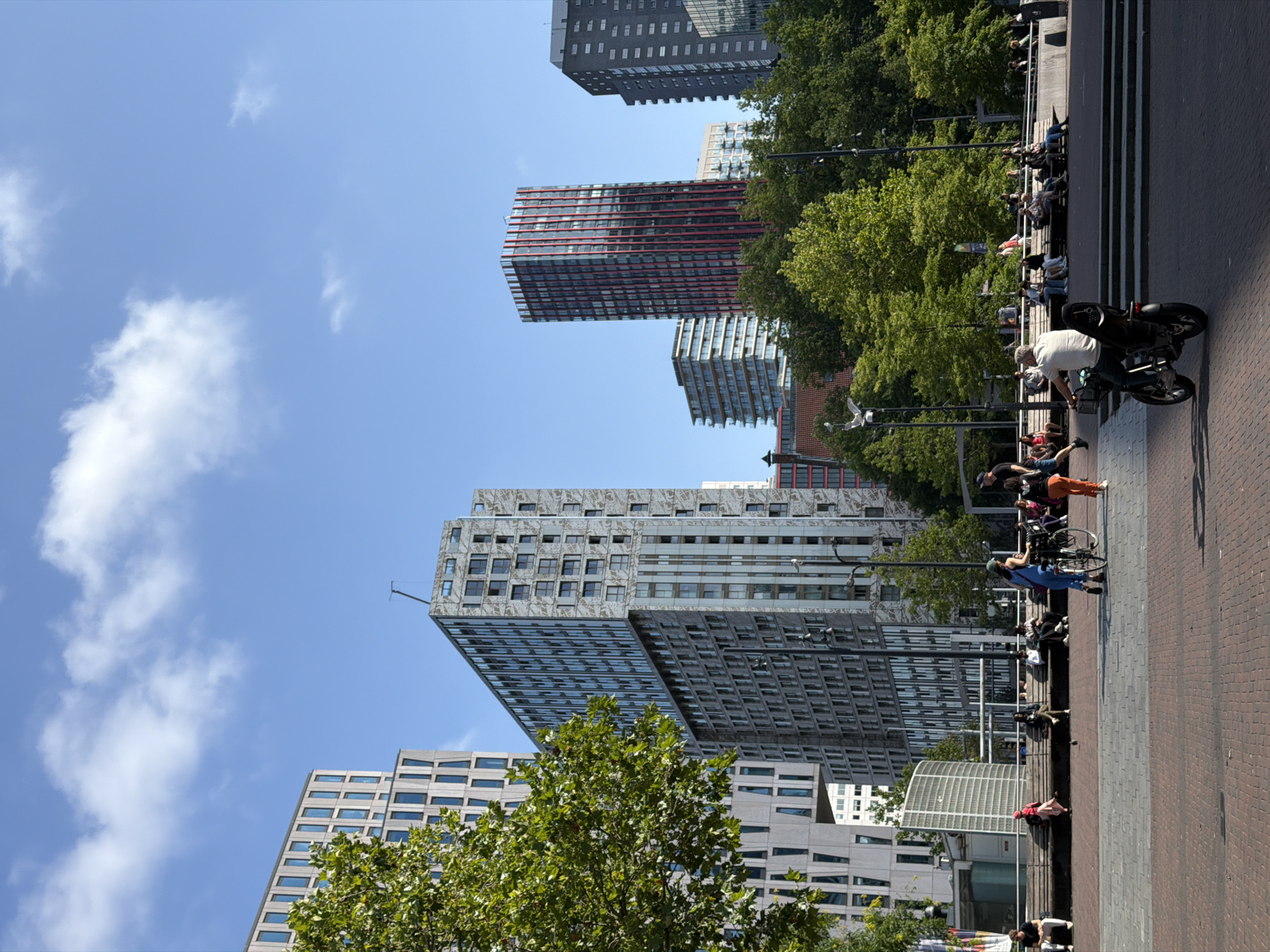 The image captures a sunny day in an urban plaza in Rotterdam, Netherlands. Modern, architecturally diverse buildings dominate the background, featuring a mix of glass facades, concrete, and decorative panels. One prominent tall building displays a striking red and white vertical striped pattern. In the foreground, people are seen relaxing on benches and walking through the paved plaza. A woman is walking a bicycle, and a man is riding a scooter. Lush green trees provide shade and visual contrast to the urban landscape. The sky is bright blue with scattered white clouds. The scene conveys a sense of a lively public space in a contemporary city.