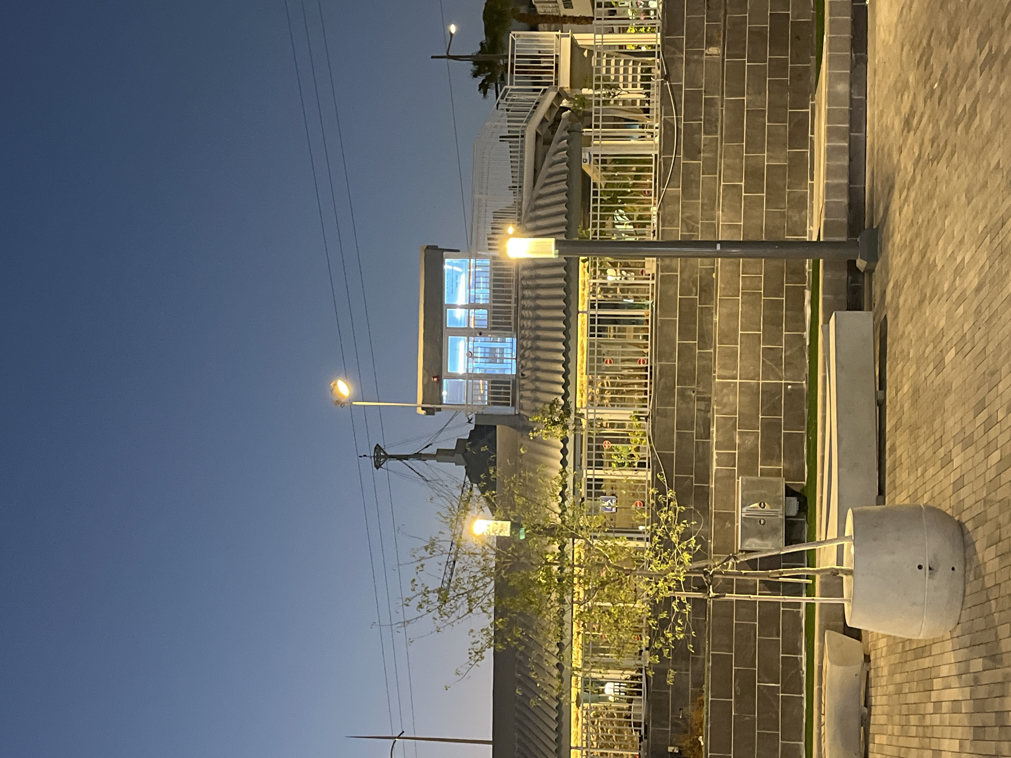 An outdoor scene at dusk shows a modern building with a flat, corrugated roof and a second-story enclosed area illuminated by cool blue lights. The building is set against a textured gray brick wall, with a white metal railing extending across its facade. In front of the wall, a small tree in a concrete planter stands on a paved walkway. Two concrete benches are visible on the left and right sides of the image. A lamppost stands in the foreground to the right of the tree, and another lamppost is visible further back on the right. The sky above is a deep twilight blue, with power lines strung across it.