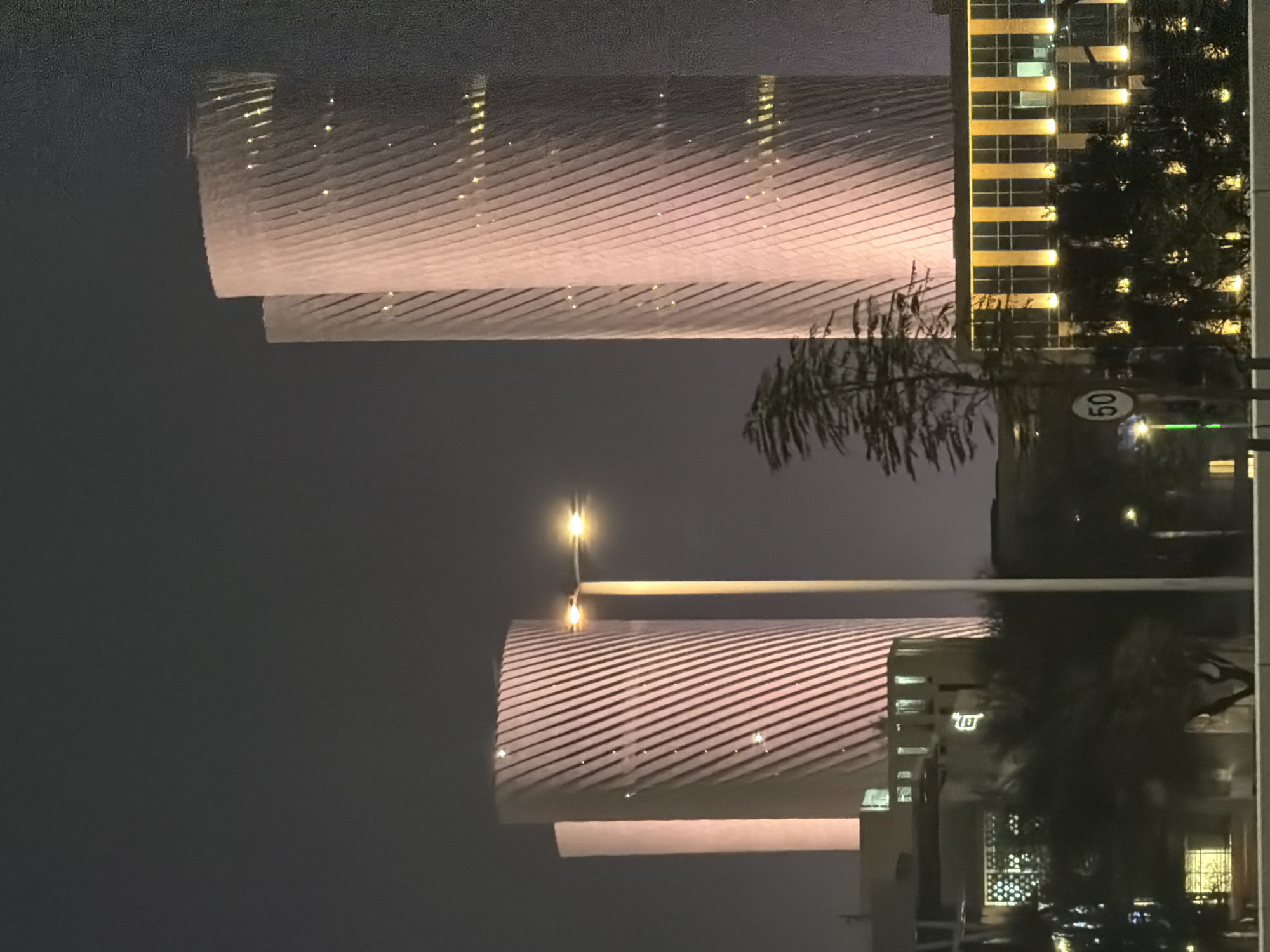 An urban nighttime scene features two illuminated, spiral-shaped skyscrapers in the background. A street lamp with two illuminated bulbs stands in the mid-ground between the skyscrapers. In the foreground, a low-rise building with a grid of yellow lights and dark trees in front of it is visible. A speed limit sign with "50" is mounted on a pole to the left of the street lamp. The sky is dark and the overall atmosphere is dimly lit.