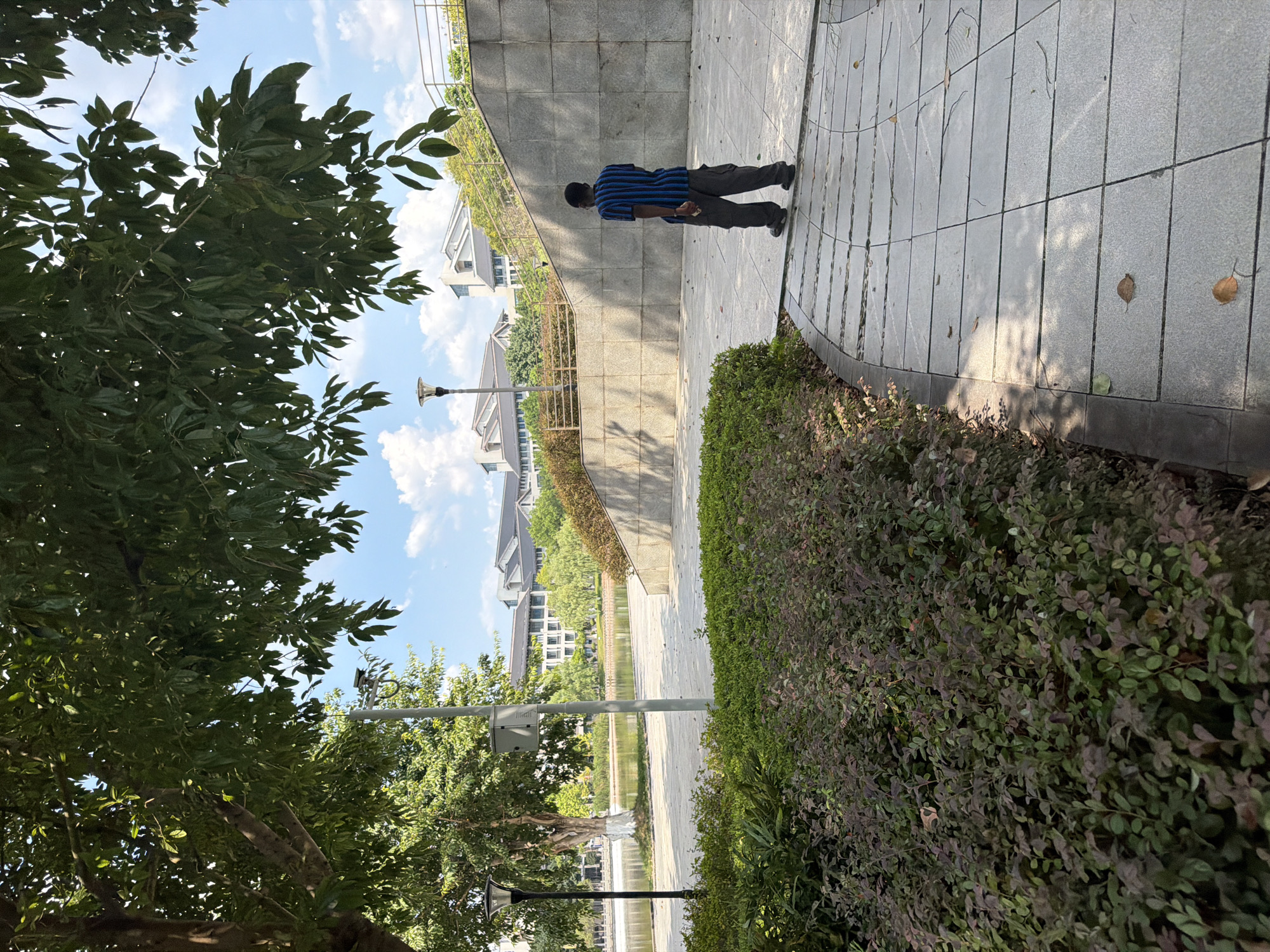 A lone Black male wearing a striped t-shirt and dark pants walks along a paved pathway in a park-like setting. The pathway curves to the right, bordered by manicured green bushes on the left and a concrete retaining wall on the right. In the background, a body of water is visible, with trees and modern buildings situated beyond it. The sky is blue with scattered white clouds. This scene is located in the city of Jiangjin, China.
