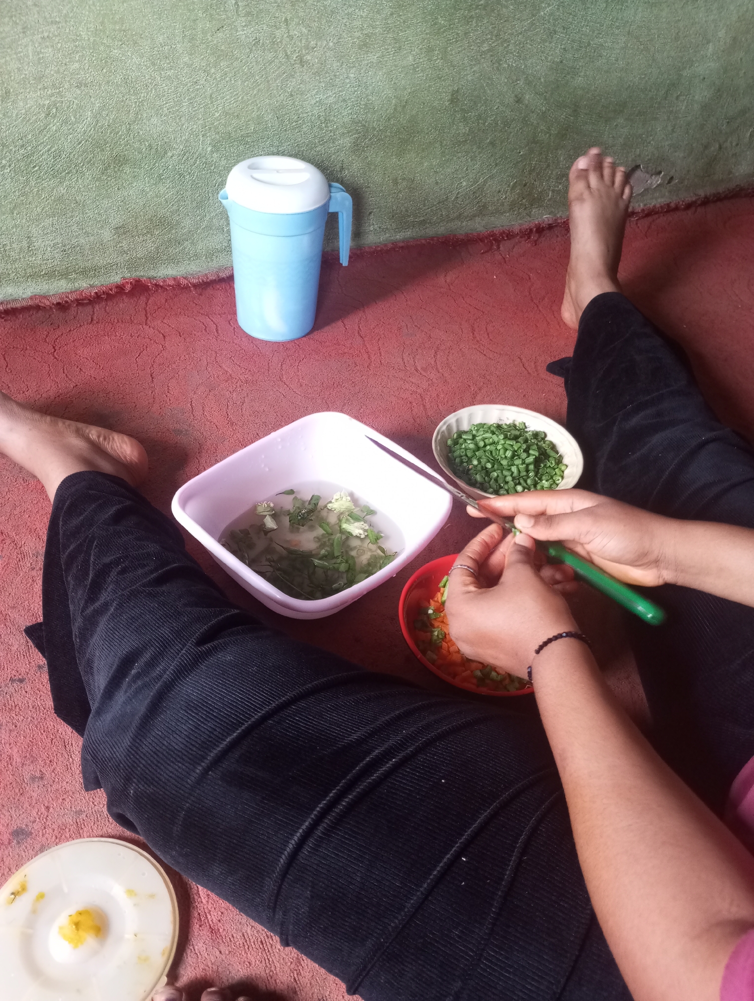 The image depicts an indoor scene on a red carpeted floor, with a textured green wall in the background. In the foreground, a person is sitting, wearing black corduroy trousers. Their legs are extended, and their feet are visible. To the left, a white plate with a yellow substance is present. In the center of the image, there are several bowls. A white square bowl contains chopped green vegetables in water. Next to it, an oval bowl holds more chopped green vegetables. A red bowl contains chopped carrots and green vegetables. A person's hands are actively engaged in preparing food, holding a green-handled knife and a piece of green vegetable. Their right hand is in the process of chopping, and they are wearing a black beaded bracelet. The overall impression is that of food preparation, possibly for a meal. The image does not contain any explicit indicators to determine its location within the city of Onitsha, Nigeria.