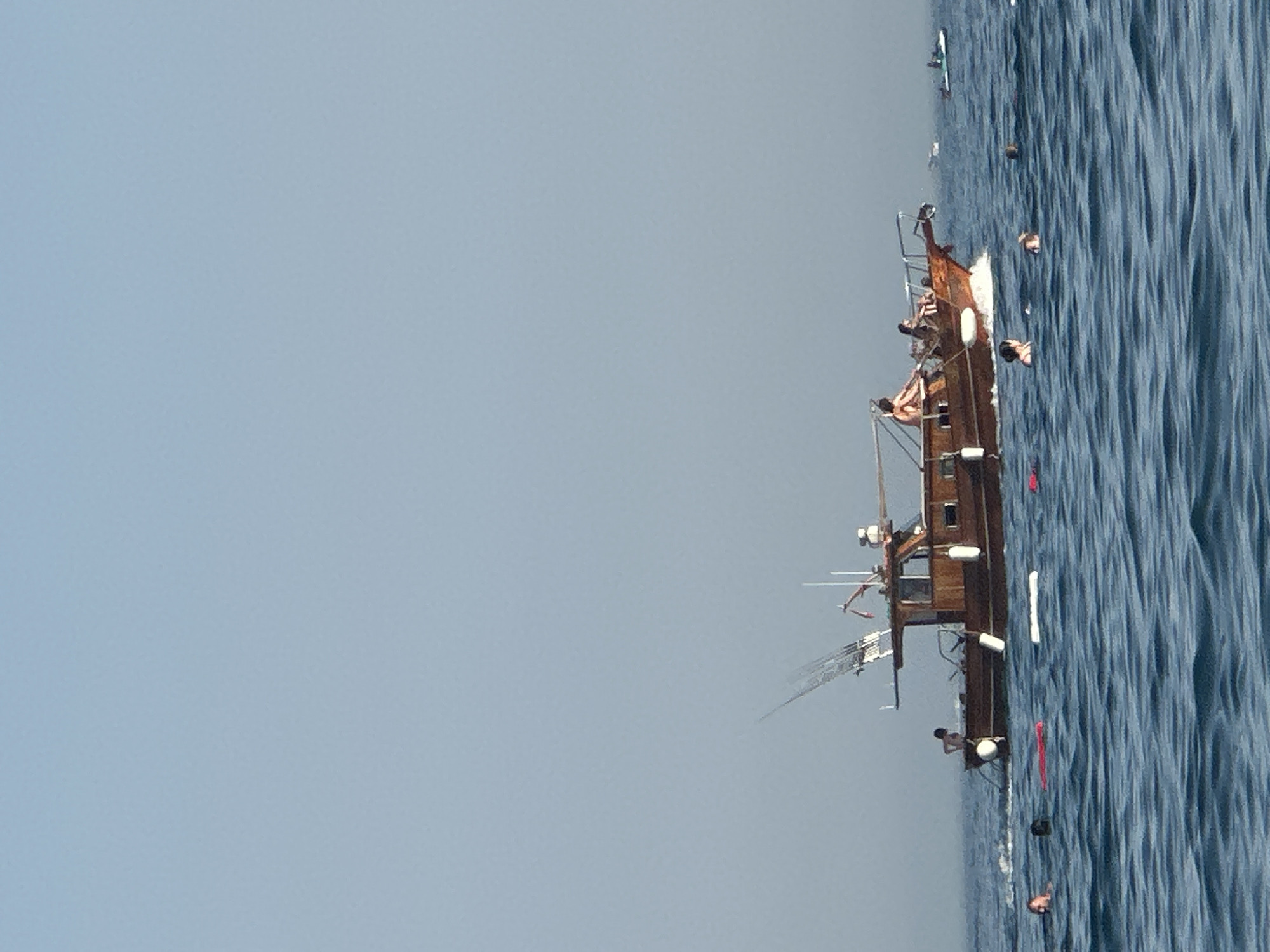 The image depicts a wooden boat with several people on its deck, some of whom appear to be fishing. The boat is moving through blue water, creating a wake. In the foreground, several individuals are swimming or floating in the water, some with inflatable aids. In the background, the sky is clear and light blue, with a faint haze visible on the horizon. The scene appears to be a leisurely day at sea, possibly near the town of Boreti in Montenegro, a coastal area known for its beaches and marine activities.