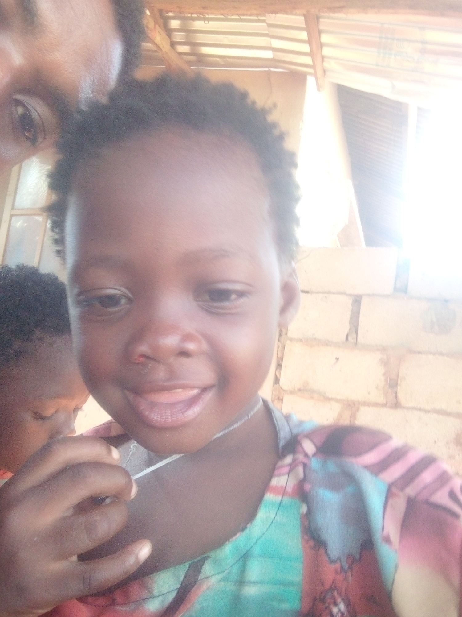 The image is a close-up, somewhat blurry photograph of a young child with dark skin. The child is smiling slightly, with their tongue visible. They are wearing a colorful shirt with shades of pink, blue, and red. A thin silver chain is visible around their neck. In the upper left corner, a portion of an adult's face is visible. Another child is partially visible on the left side, with their head turned away from the camera. The background includes what appears to be a brick wall and a corrugated metal roof. The location is Unknown, Unknown.