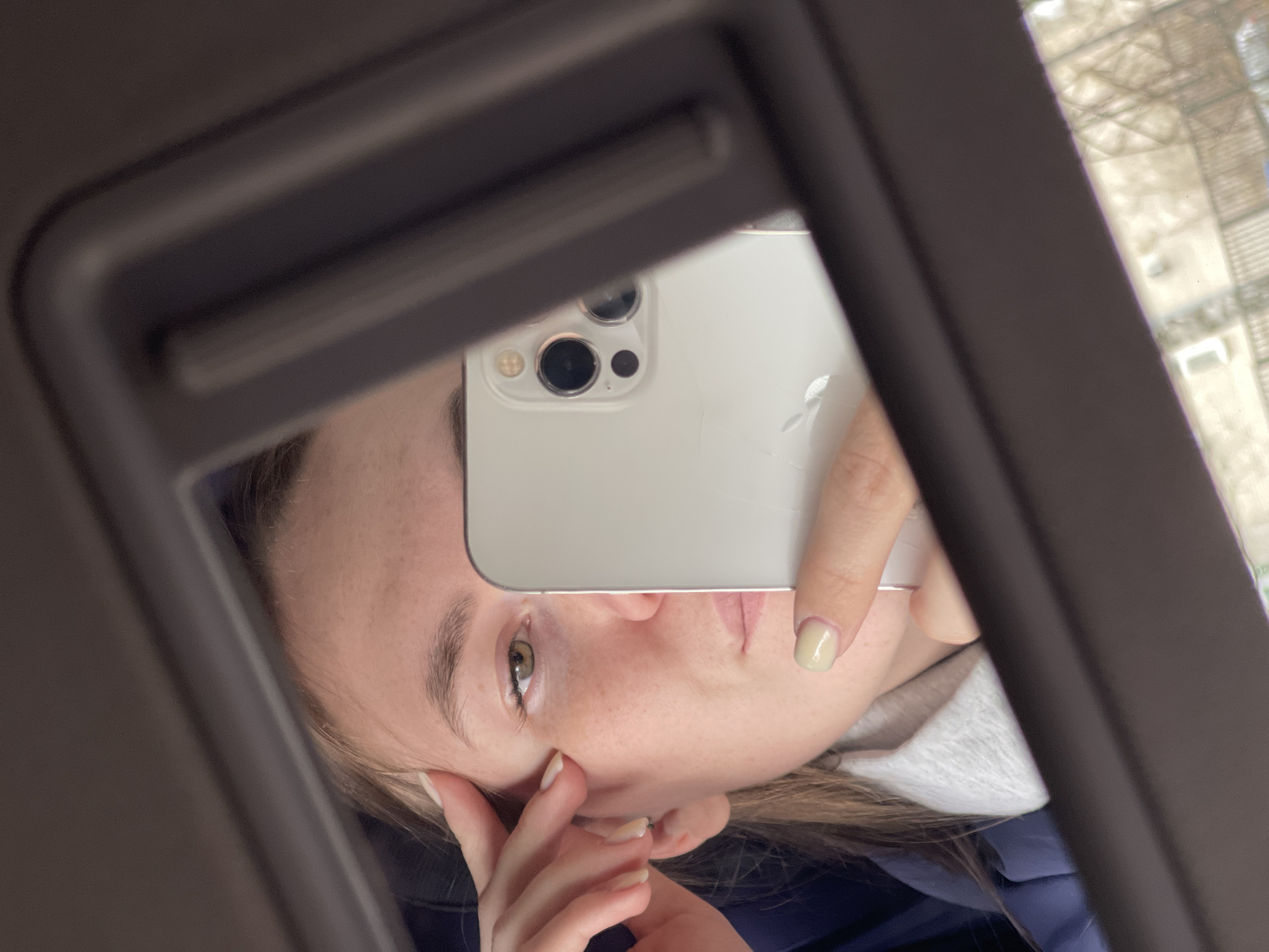 This media file captures a reflection selfie of a young person, likely female, within what appears to be an enclosed space, possibly a vehicle. The image is oriented vertically, but the reflection shows the person's face rotated sideways.
The subject has fair skin, light brown hair, and a visible green or hazel eye. Her expression is neutral. One hand holds a light-colored smartphone, identifiable as an iPhone due to its distinctive triple camera setup on the back. The Apple logo on the phone's back appears to have a crack or stylized damage. The thumb and index finger holding the phone have pale yellow nail polish. Her other hand rests near her face, revealing natural-colored nails. She is wearing a grey top, possibly a hoodie or sweatshirt, layered over a dark blue or black garment.
The reflection is contained within a dark, possibly metallic or plastic, frame, suggesting a mirror, window, or screen inside a car, bus, or similar interior. Beyond the reflected frame, a blurred background depicts what seems to be a building or structure with a grid-like pattern, indicating an outdoor, possibly urban, environment under daylight.
The activity taking place is the act of taking a mirror selfie or a reflection photograph with the smartphone. No visible text is present in the image. Given the context, the location is Chaikovsky, Russia.