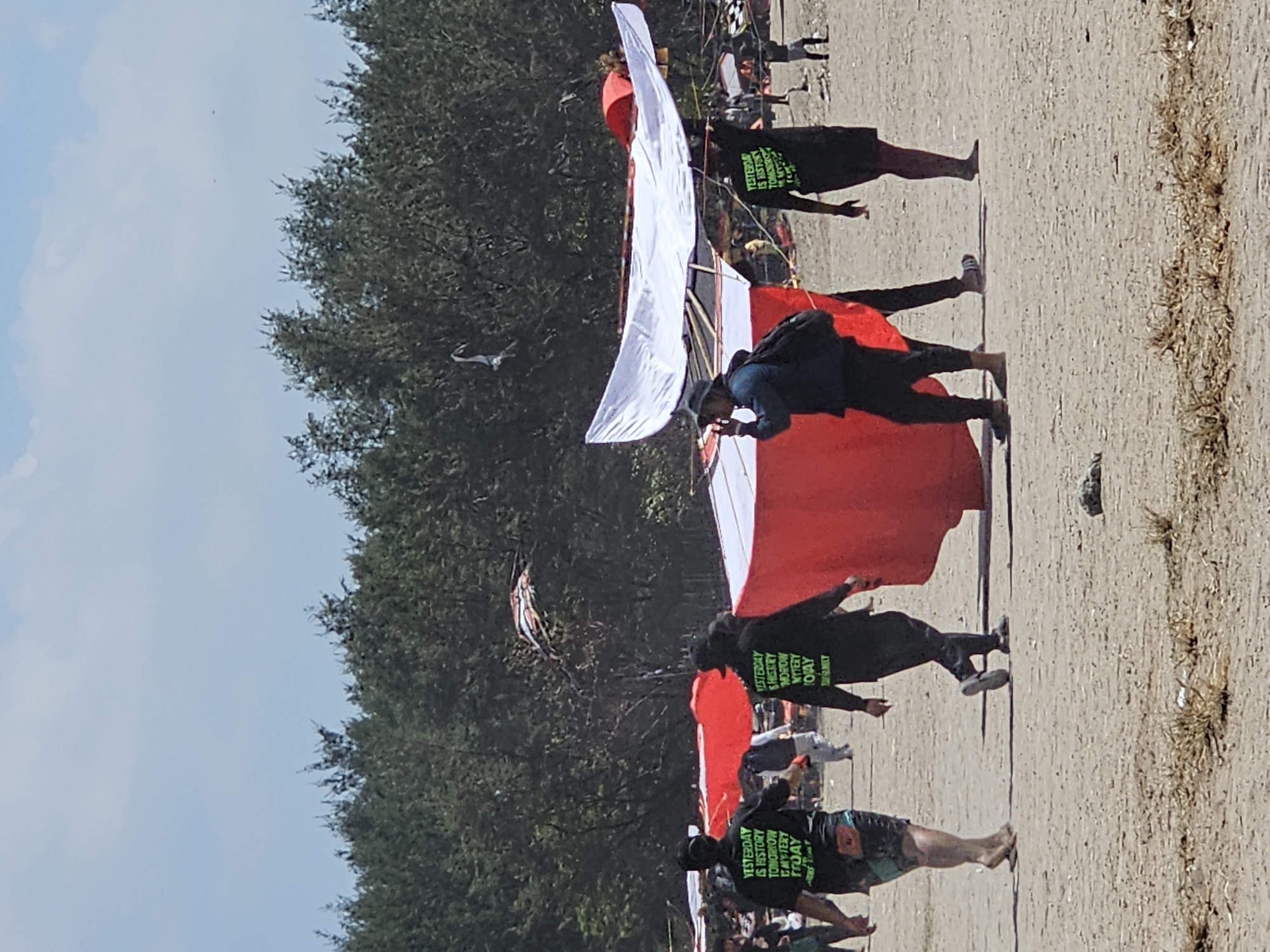 The image depicts a group of people on a sandy beach with a line of trees in the background. Several individuals are walking, carrying or assembling large, colorful kites. Two individuals in the foreground are wearing black t-shirts with green text that reads "YESTERDAY IS HISTORY TOMORROW IS MYSTERY TODAY IS FAMILY." One person is wearing a hat and holding a phone to their ear. The kites appear to be elaborate structures, with one prominently featuring white and red fabric. The environment suggests a sunny day with a clear sky, hinting at a festival or event related to kite flying. The scene is consistent with popular kite festivals held annually in Denpasar, Indonesia, such as the Denpasar Kite Festival which typically takes place around June or July.
