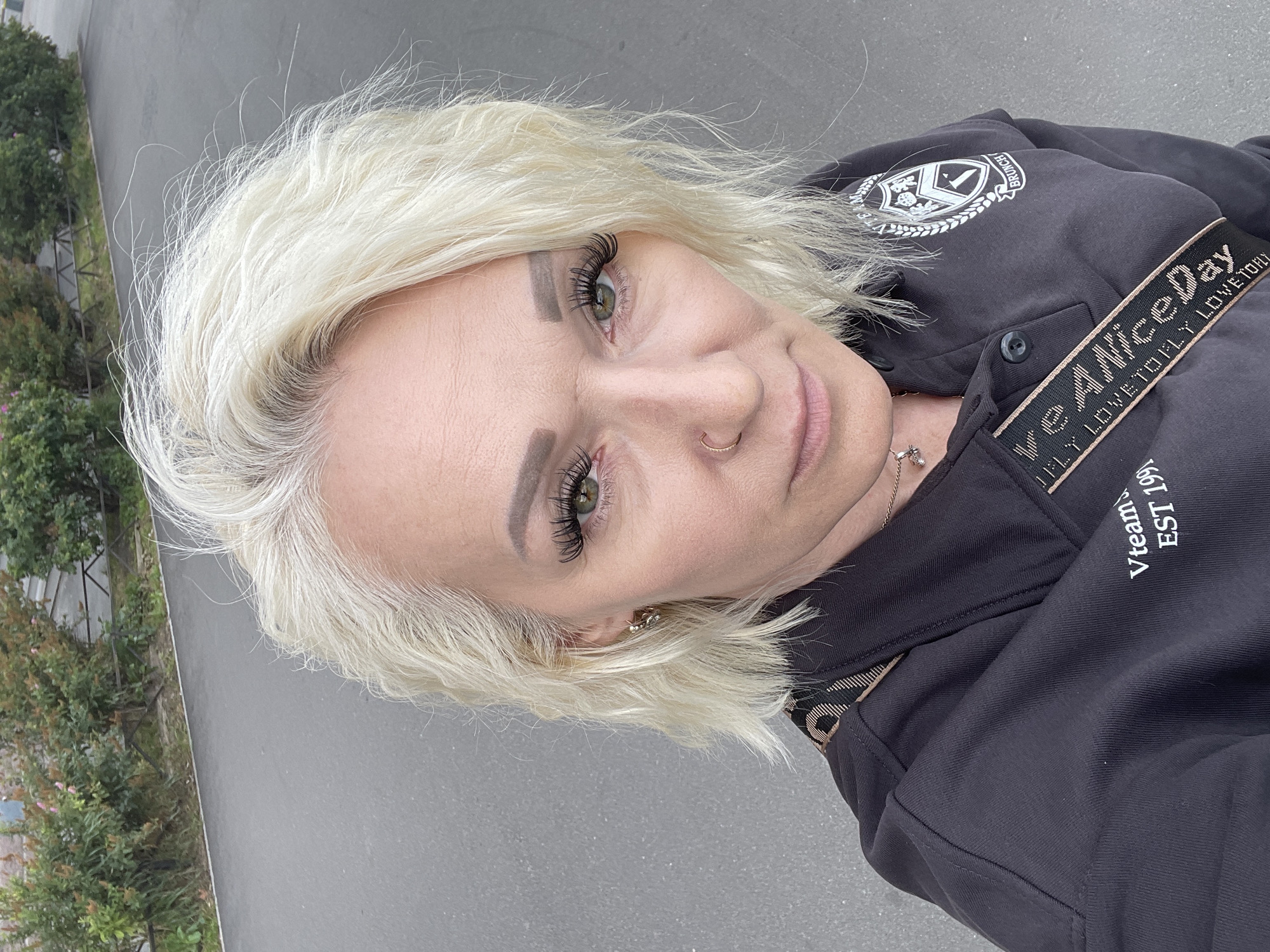 A close-up, high-angle outdoor shot shows a fair-skinned woman with blonde, wavy hair and green eyes. She is looking directly at the camera with a neutral expression. She has prominent eyelashes and wears subtle makeup, including eyebrow pencil and lip gloss. A gold nose ring is visible on her right nostril, and a small, gold pendant hangs from a delicate chain around her neck. She is wearing a dark grey polo shirt with white text and a crest on the left side of the chest. A black strap with gold text runs diagonally across the shirt from her left shoulder to her right side. The background consists of a grey asphalt surface, a curb, and green bushes behind a black metal fence. The lighting suggests an overcast day.