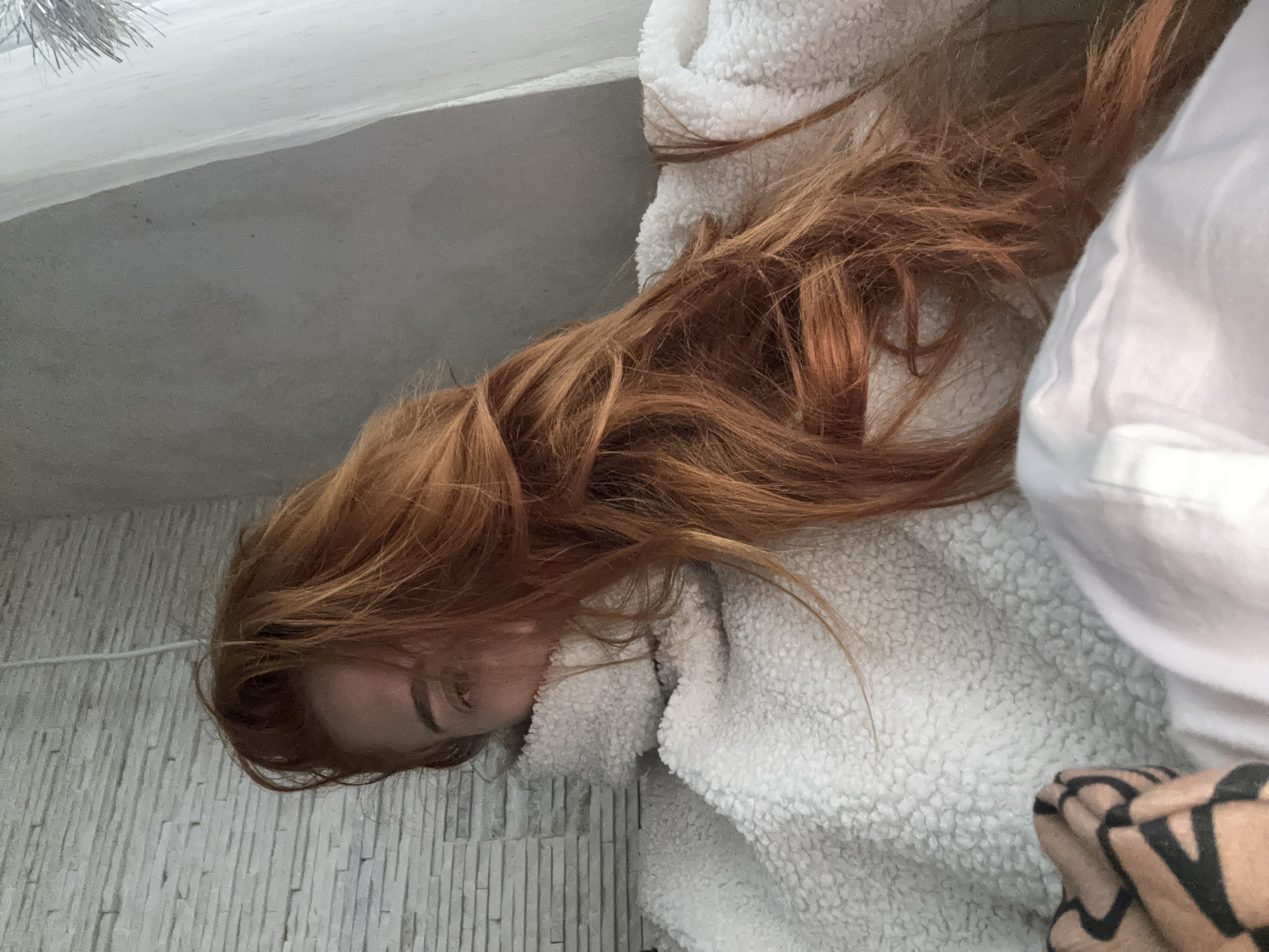 A red-haired woman is lying down, presumably inside a room. The walls are light gray with a vertical texture resembling stone or stacked tiles. She is wearing a white, fluffy jacket or blanket. A portion of patterned clothing or a blanket is visible at the bottom right corner.