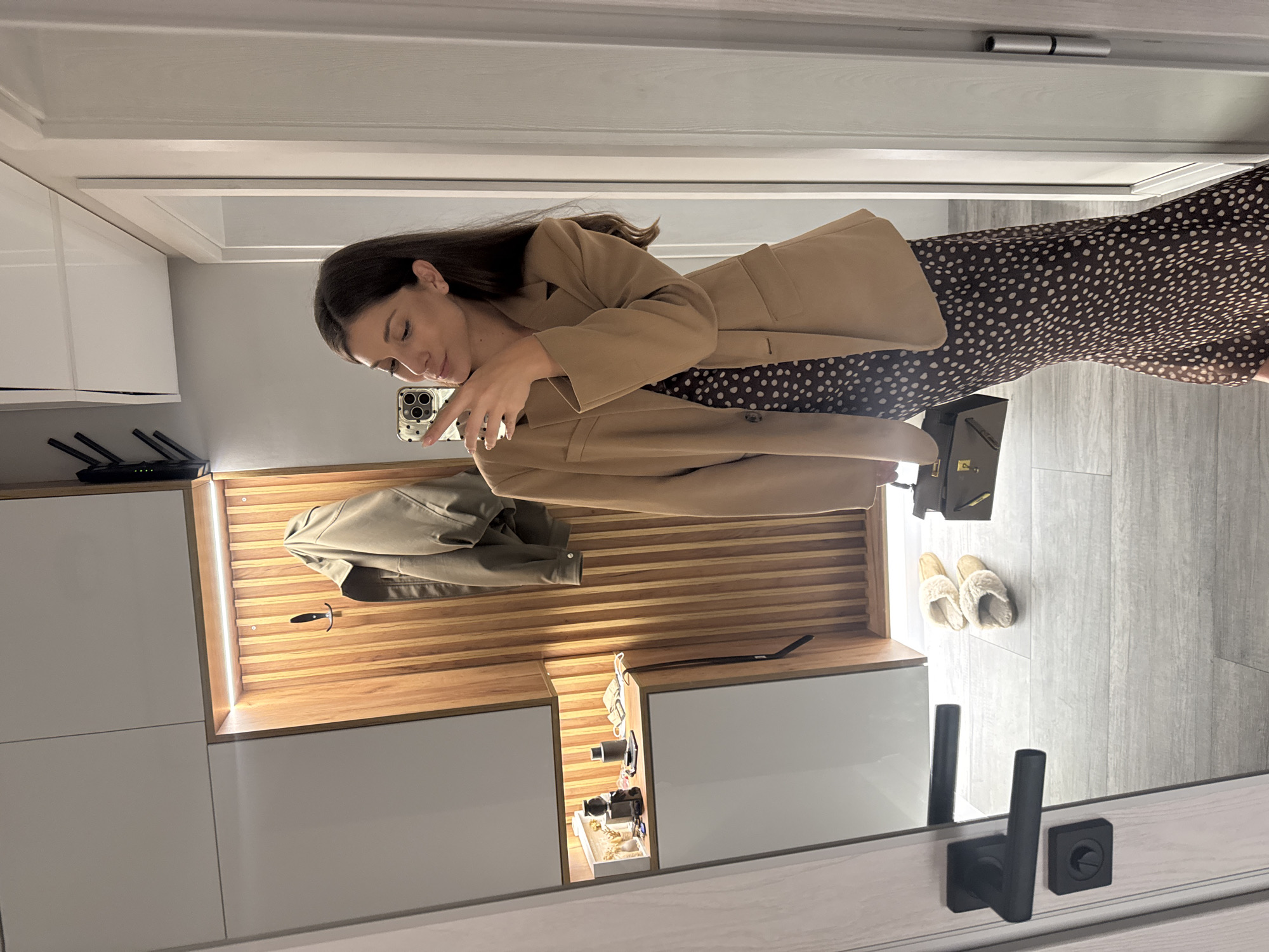 A woman is taking a selfie in a mirror indoors. She is wearing a brown blazer and a brown and white polka dot dress. Behind her is a wall unit with white cabinets and a wooden shelf. On the shelf is a tan jacket and a black hook. Below the shelf is an open compartment with various small items. On the floor, there is a black bag and a pair of fuzzy slippers. The floor is gray tile. The reflection in the mirror suggests that the photo was taken in a hallway or entrance area. The location in the city of Burgas, Bulgaria, cannot be determined from this image.