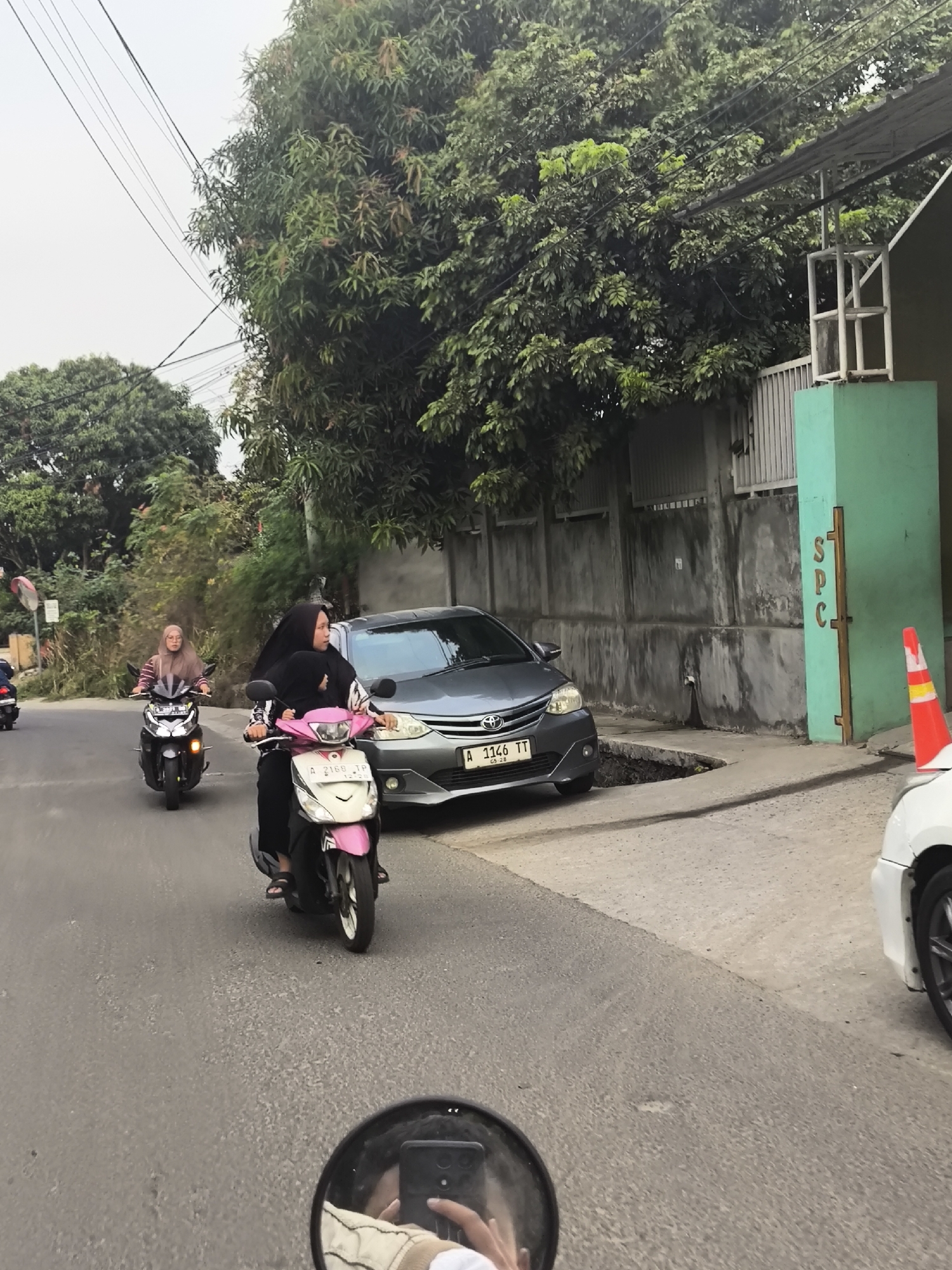 The image captures a street scene, possibly in Cilegon, Indonesia, with several motorcycles and a car visible. In the foreground, a person is riding a pink and white scooter, with another person riding pillion, both wearing hijabs. To their left, another person on a black scooter, also wearing a hijab, is riding ahead. A gray car is parked on the right side of the road, next to a concrete wall and a green pillar. The street appears to be paved, and there are trees and power lines overhead. A traffic cone is also visible near the parked car. The perspective suggests the photo is taken from the viewpoint of someone riding a motorcycle.