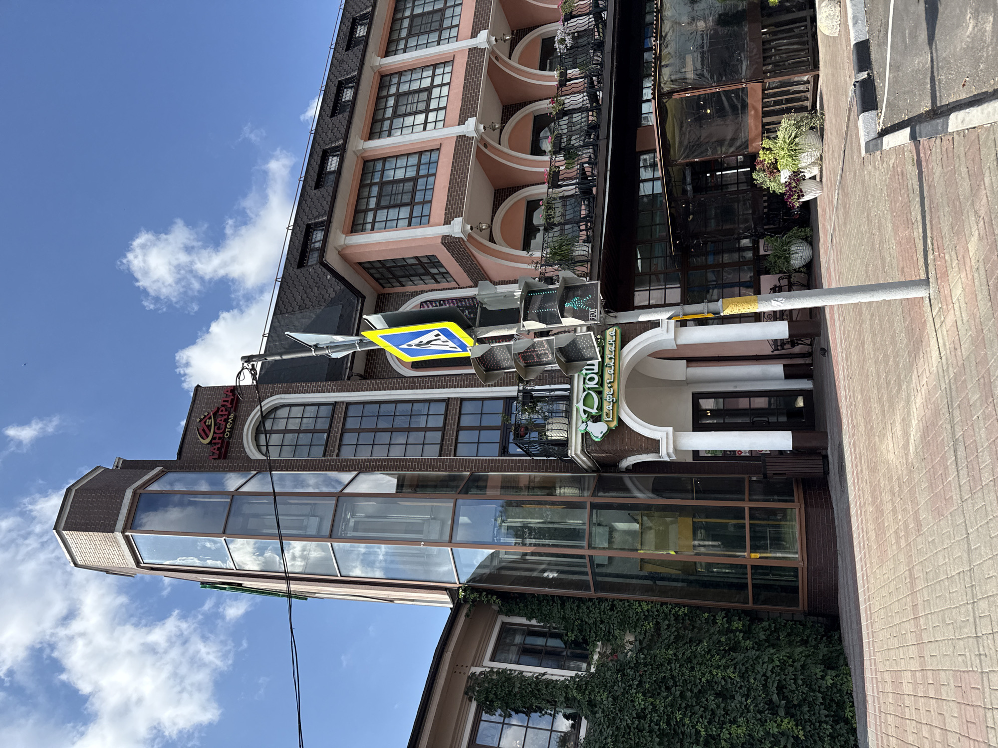 This is a medium-shot, eye-level, outdoor, daytime photograph of a multi-story building located in Tomilino, Russia. The building features a modern glass elevator shaft on the left, an arched entrance with a canopy, and a pink upper story with numerous windows. The architecture is a mix of brick and pastel colors, with a dark-tiled roof. A pedestrian crossing sign and traffic lights are visible in the foreground, partially obscuring the building's entrance. The sidewalk is paved with brick, and an asphalt parking area is present to the right. Green ivy covers a portion of the building's left facade. The sky is blue with scattered clouds. There are no people or animals clearly visible in the image.