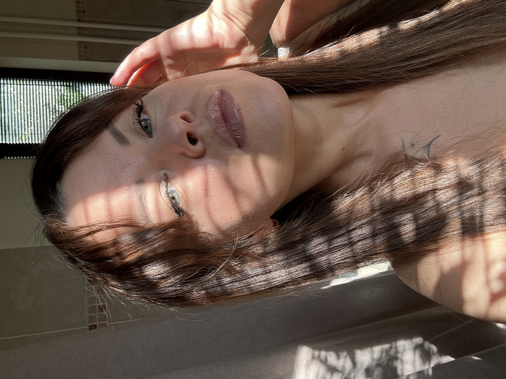 A woman with long brown hair and light blue eyes is looking towards the camera, her face partially obscured by a shadow from blinds. She is wearing light-colored makeup, including lipstick. Her right hand is raised to her temple, with her fingers splayed. A small tattoo is visible on her chest. The background appears to be indoors, with a curtain on the left and blinds with a view of greenery on the right. The lighting suggests it is daytime, with sunlight casting shadows.