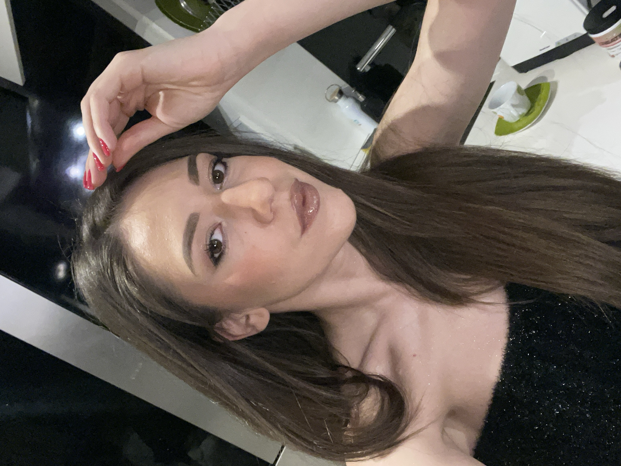A woman with long brown hair is taking a selfie. She is wearing a black sparkly top. Her left arm is raised with her hand near her forehead. She has red nail polish. In the background, there is a white countertop with various items, including a cup and saucer and a water bottle. The background is a black and white tiled wall. The location is most likely inside a home, and no information suggests that the scene is specifically located in Sofia, Bulgaria.