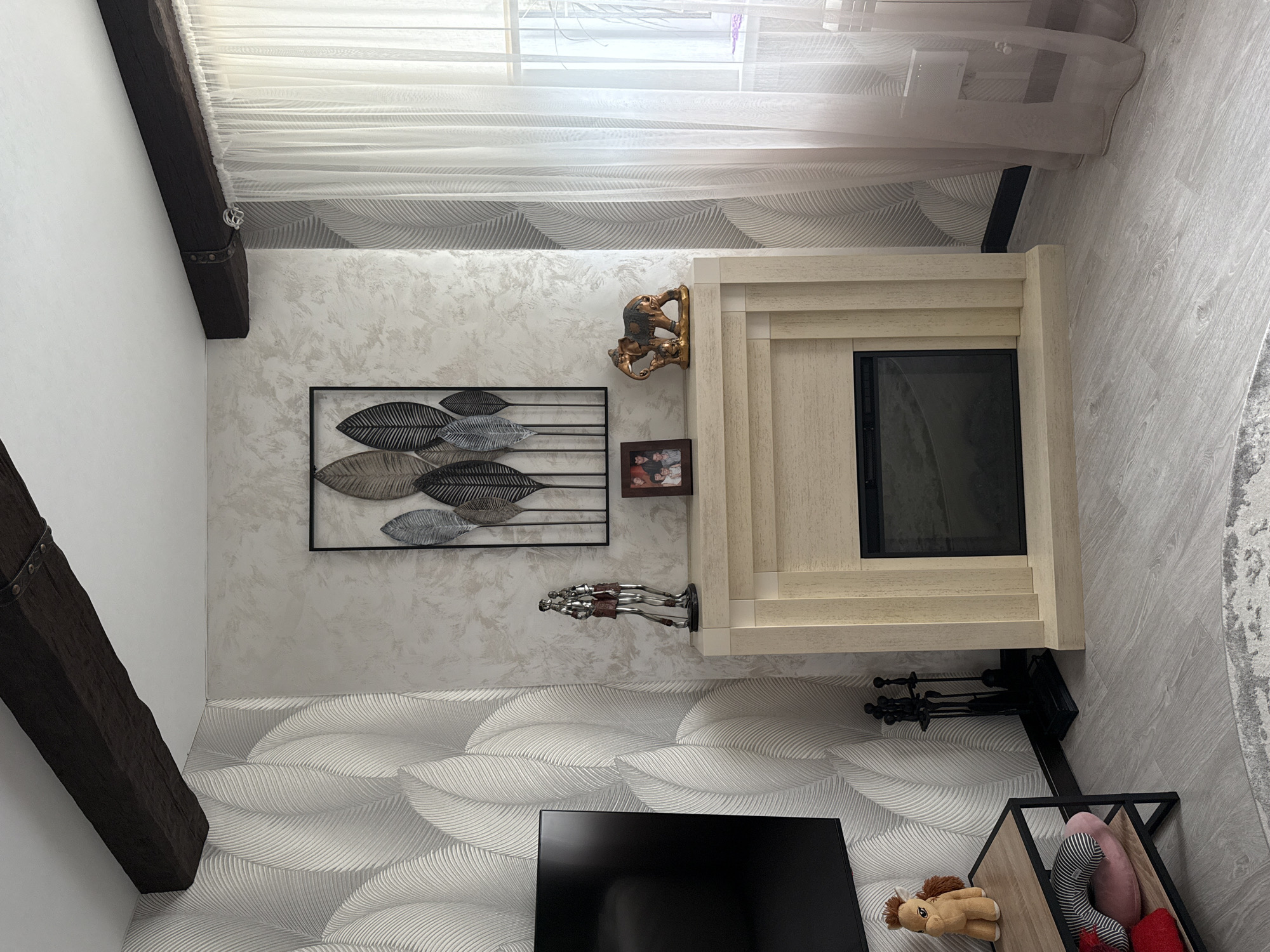 This media file captures an interior scene, likely a living room, with a modern and cozy aesthetic. The room features light-colored walls, some adorned with a subtle textured or patterned wallpaper, and a light wood-look flooring. An exposed dark wooden beam runs across the ceiling and down a wall, adding a rustic touch.
A prominent feature is a light-colored, possibly wooden, fireplace mantel with a dark, modern insert. To the left of the fireplace, on the textured wall, hangs a large black metal wall art piece depicting stylized leaves in varying shades of grey and silver. Below this, a small framed photograph is visible. Above the fireplace mantel, a golden decorative figurine, possibly depicting elephants or monkeys, is perched. Another decorative piece featuring two silver knight-like figures stands on the mantel to the left of the fireplace opening.
Below the fireplace, black metal fireplace tools are neatly stored. To the left, a television screen is visible, angled towards the viewer. A low storage unit with a wooden base and black metal frame is positioned to the right of the TV, holding several plush toys including a brown horse, a striped animal, and a red heart-shaped item.
Natural light filters into the room through a window covered by sheer white curtains, suggesting it is daytime. The flooring to the right of the fireplace is a light wood-look material, and a patterned area rug is partially visible on the far right. The wallpaper has a distinct wavy or leaf-like pattern, creating a sense of movement.
The scene is static, with no activity taking place, and no discernible text is visible on any objects. The setting is an interior space in Krapivka, Russia.