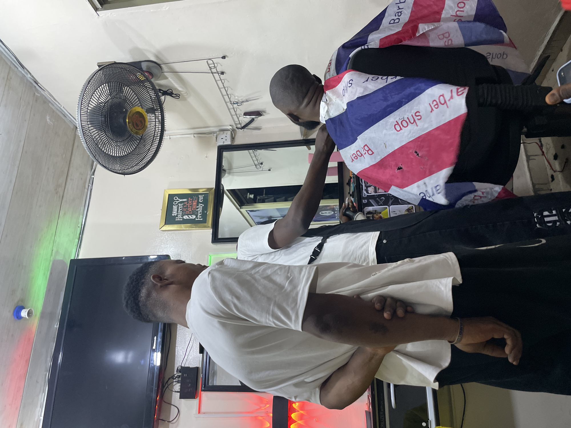 The image shows a scene inside a barber shop in Ado, Nigeria. A barber is cutting a client's hair. The barber wears a white t-shirt and the client is covered with a barber cape that has "Barber Shop" printed on it. The client is wearing black clothing. The shop features a wall-mounted fan, a mirror, a television, and a sign that reads, "SHAVE Haircut is a Barber Freshly cut". There is also a sterilizer in the shop. The walls are painted in a light color.