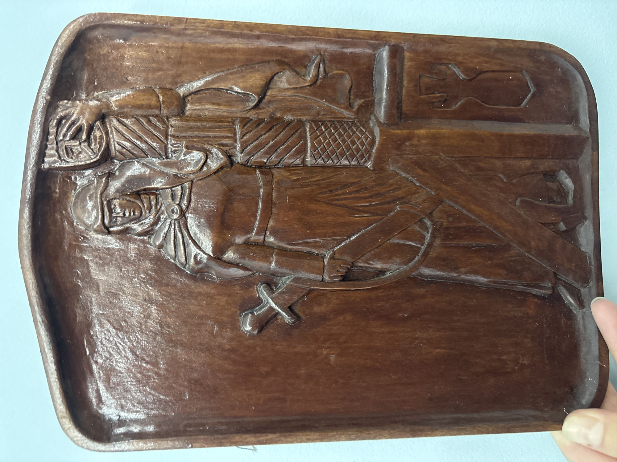 This is a close-up, rotated shot of a carved wooden tray, likely a decorative piece or a shallow dish. The carving depicts a figure, possibly a knight or a soldier, in profile. The figure wears a helmet, a tunic with textured armor detailing on the chest and shoulders, and appears to be holding a sword at their side. The sword is small and stylized, with a crossguard. The background of the carving shows what could be a backdrop or part of a scene, with some geometric patterns and what appears to be a cross-like symbol at the top right. The wood is a dark brown, with a polished finish that highlights the carved details and the grain of the wood. The tray has a slightly raised, rounded edge. The lighting is bright, casting subtle shadows within the carvings, emphasizing their depth. The image is taken from a slightly overhead angle. The location context provided is Novaya Usman', Russia, but there are no explicit visual cues within the image to confirm or deny this. There is no visible text in the image.