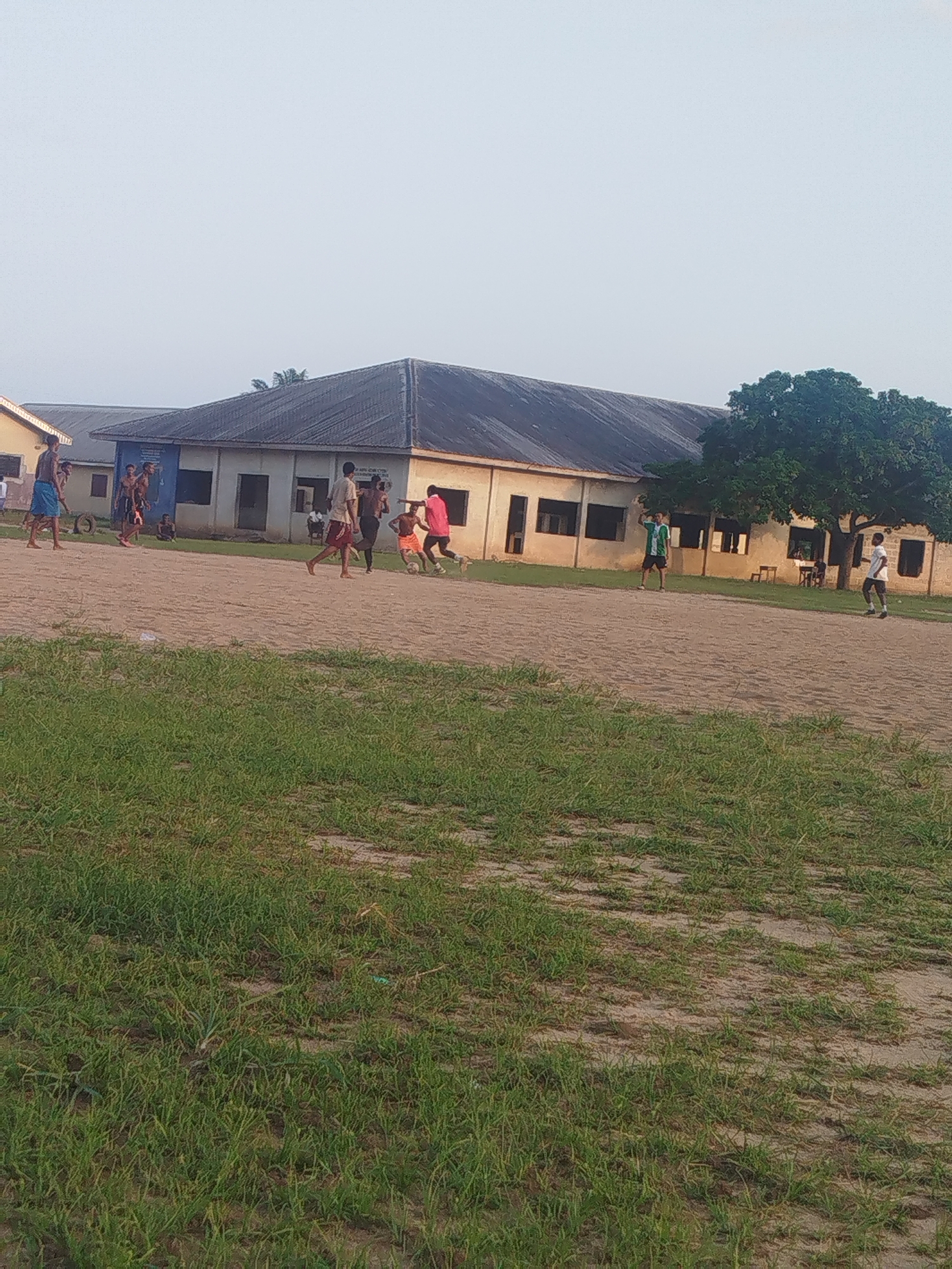 The image depicts a group of young people playing soccer on a sandy field in Ikot Mbonde, Nigeria. In the background, there is a low, long building with a dark roof and light-colored walls. A tree stands to the right of the building. The foreground is covered in green grass with sandy patches. The overall lighting suggests it is either early morning or late afternoon. Several players are actively engaged in the game, while others stand by observing. The scene appears to be a casual, informal soccer match.