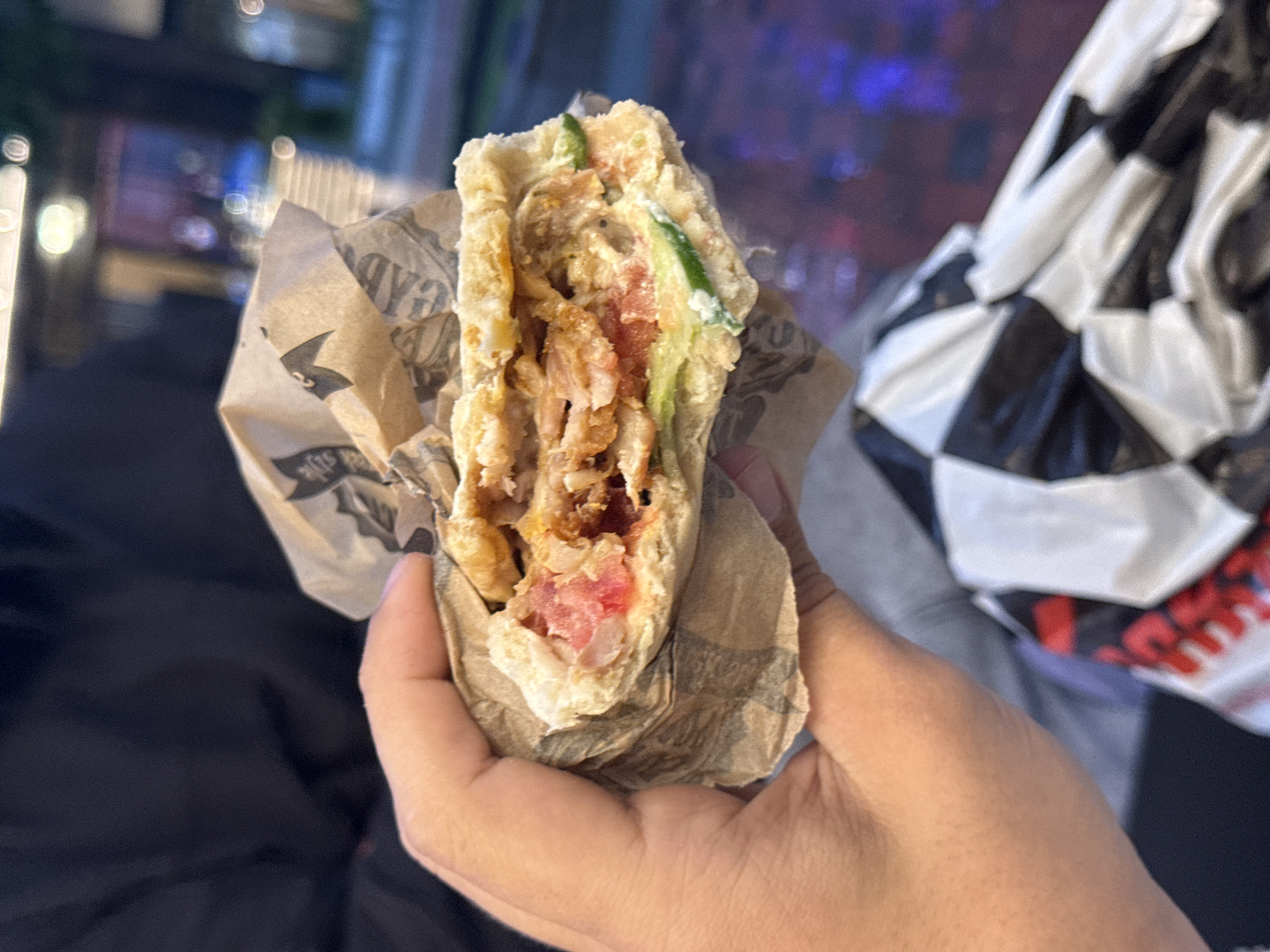 The image features a close-up of a person's hand holding a street food item, likely a wrap or kebab, cut in half to display its internal components. The filling consists of shredded, cooked meat (possibly chicken), bright red diced tomatoes, slender green vegetable strips (such as cucumber), and a visible creamy white sauce. The food is partially contained within a light brown paper wrapper, which shows faint, stylized black text, including characters like "AD" and "YS," along with a small, indistinct logo that resembles a crown or abstract symbol.
The setting appears to be outdoors or in a dimly lit urban environment at night, indicated by the out-of-focus, circular lights creating a bokeh effect in the background. To the right of the hand, a prominent black and white checkered pattern is visible, which could be part of a bag, jacket, or another object. Dark, plain fabric is also present in the lower left of the frame. The activity depicted is simply holding the food, suggesting the person is about to eat or has just started eating it. No specific emotions are discernible, and weather conditions cannot be determined. The image portrays a casual dining experience in what is understood to be Varna, Bulgaria.