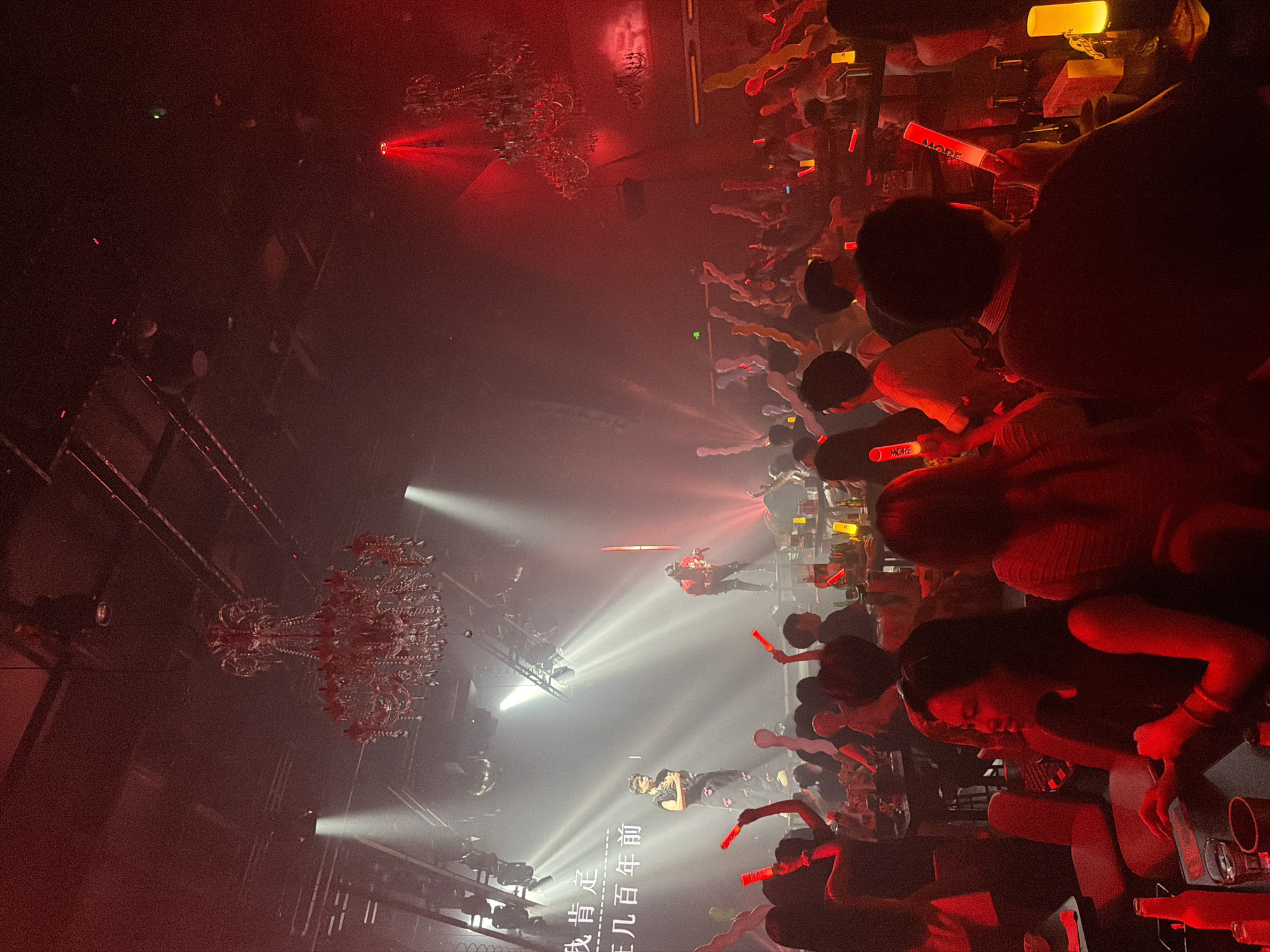 The image depicts an indoor entertainment venue, likely a nightclub or performance space, located in 江北城街道, China. The scene is illuminated predominantly by red light and dense atmospheric haze.
In the central background, a stage features two male performers. The performer on the left stands centrally, wearing a dark, patterned sleeveless top and dark trousers, with hands clasped near his chest. To the right, another performer stands on a raised platform, clad in a dark and red jacket, holding a microphone. Behind them, a large screen displays white Chinese characters: "我肯定" (Wǒ kěndìng) and "早几百年前" (Zǎo jǐ bǎi nián qián). Multiple white spotlights project beams across the stage area, cutting through the haze. Several disco balls are suspended from the ceiling above the stage. A tall, slender red light fixture stands vertically to the right of the performers.
Two ornate, crystal-like chandeliers, heavily illuminated with red light, hang from the ceiling. One is prominent in the mid-left, and another smaller one is visible further to the right. The ceiling structure reveals exposed dark beams and lighting rigs.
The foreground and midground are occupied by a large audience, gathered at tables and standing. Many individuals are holding and waving glowing light sticks or batons; most are red, some are yellow. A few inflatable noisemakers are also visible among the audience. In the immediate foreground-left, a woman with dark hair is seated at a table, looking towards her left. On the right foreground, a person with their back to the camera holds a red light stick branded "MODE." Tables in the audience area hold bottles, glasses, and small, warm-toned table lamps. The overall impression is of an active performance event.