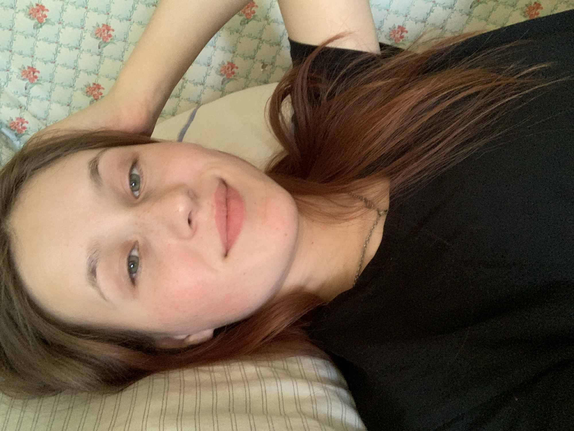 A young woman with fair skin, green eyes, and reddish-brown hair is lying down on a bed. She is wearing a black shirt and a delicate silver necklace. Her left arm is raised above her head, resting on a pillow covered with a floral patterned sheet. The bed has striped bedding. The location in the city of Фоки, Russia, is undetermined.